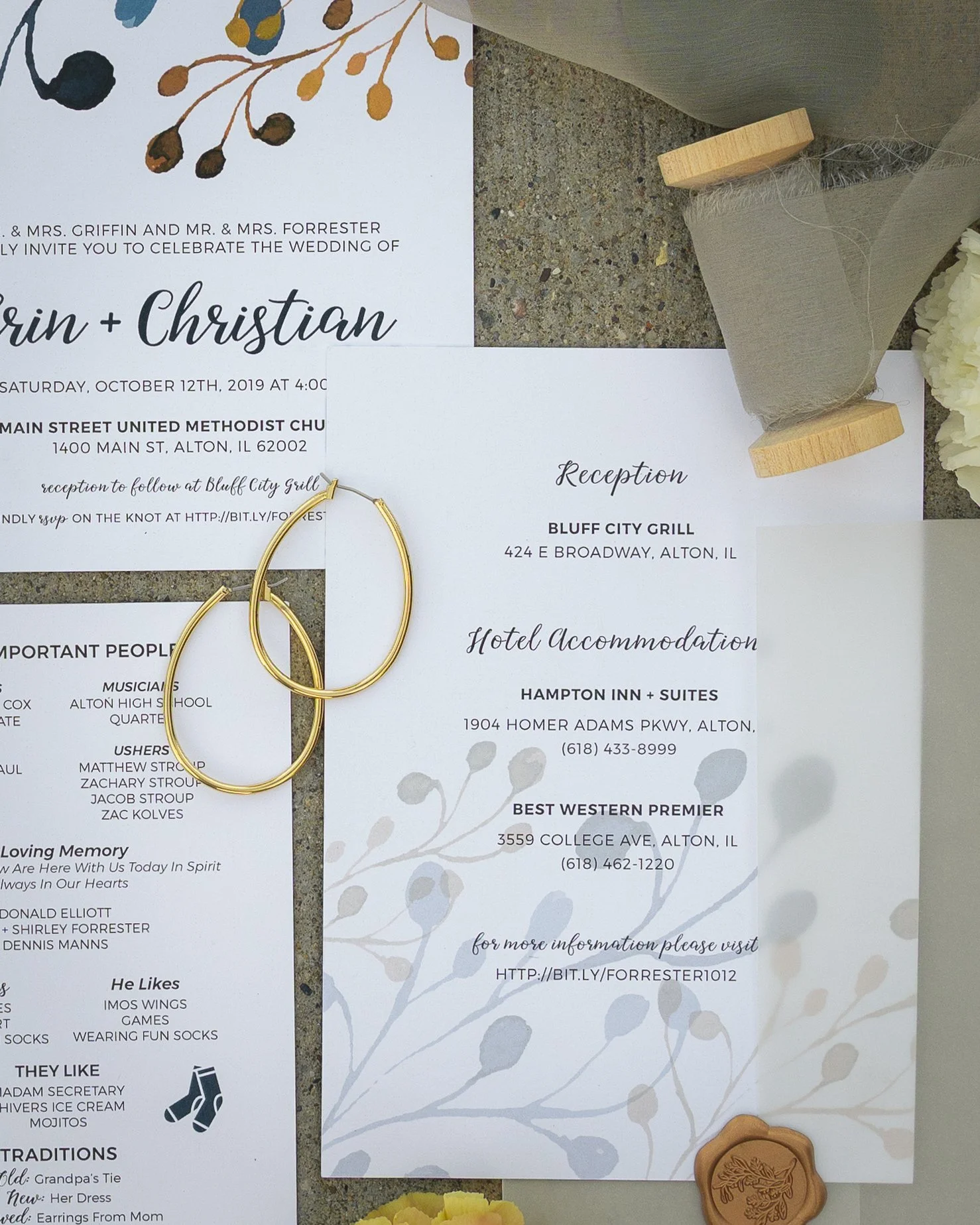 Wedding invitation and reception details for Erin and Christian's wedding at Bluff City Grill in Alton, Illinois, with gold earrings, a wooden stamp, and a floral accent on a gray surface.