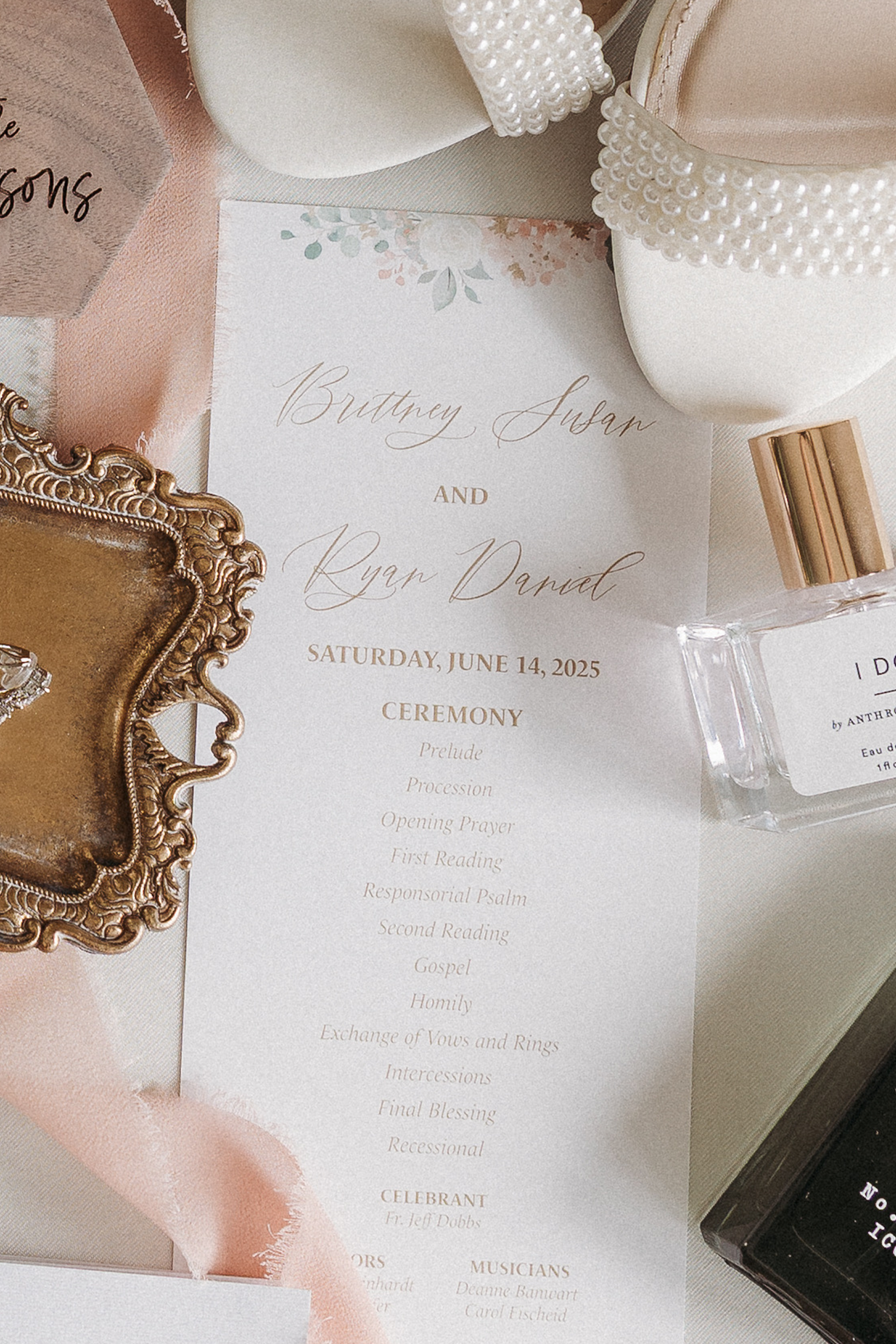 A wedding program for Brittney Susan and Ryan Dared with a floral design on top, placed on a satin fabric with various decorative items including a pearl-embellished shoe, a gold-framed mirror, and perfume bottles.