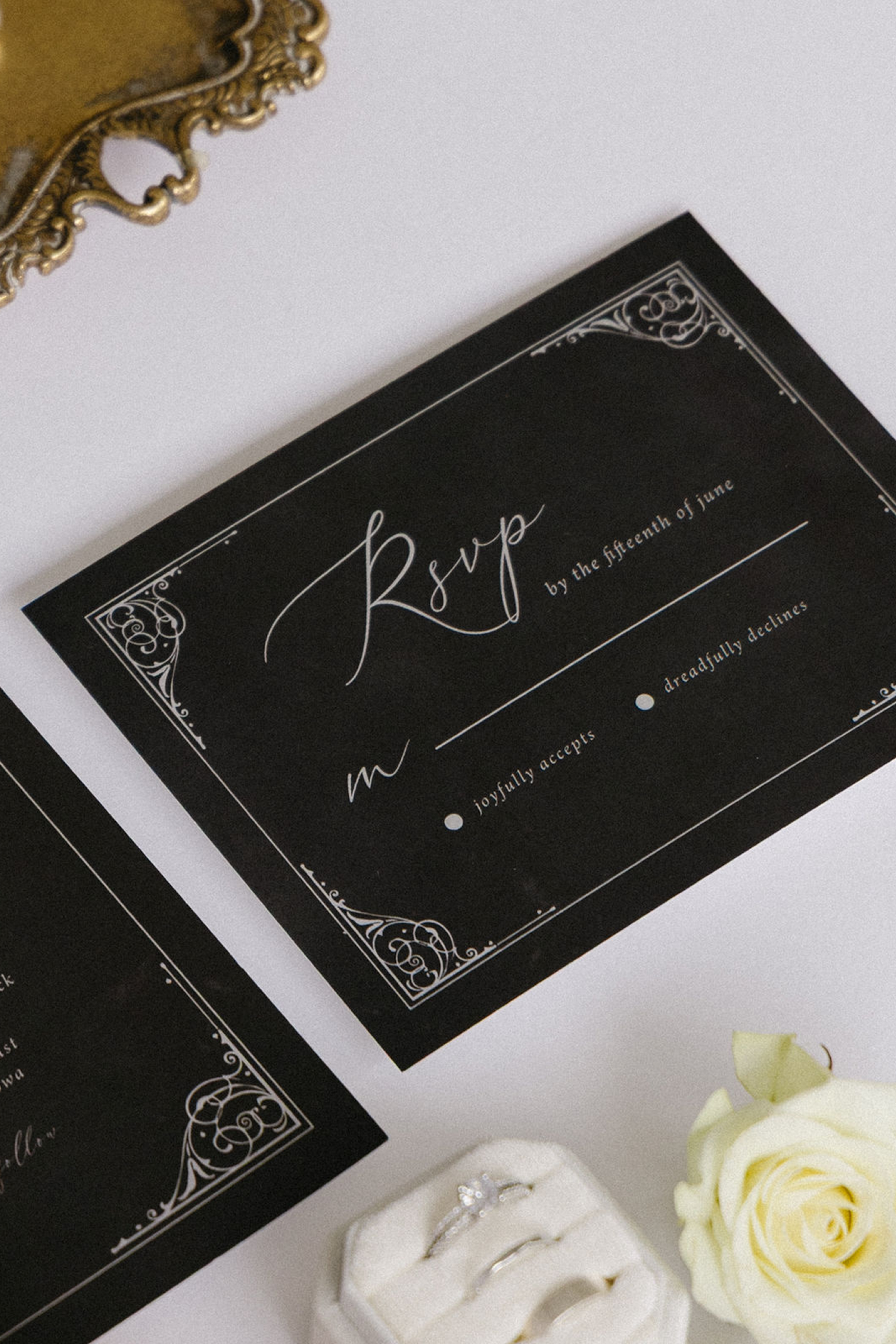 A black wedding invitation with white ornate border designs and the word 'RSVP' written in elegant script, along with a white rose and a small jewelry box containing rings.