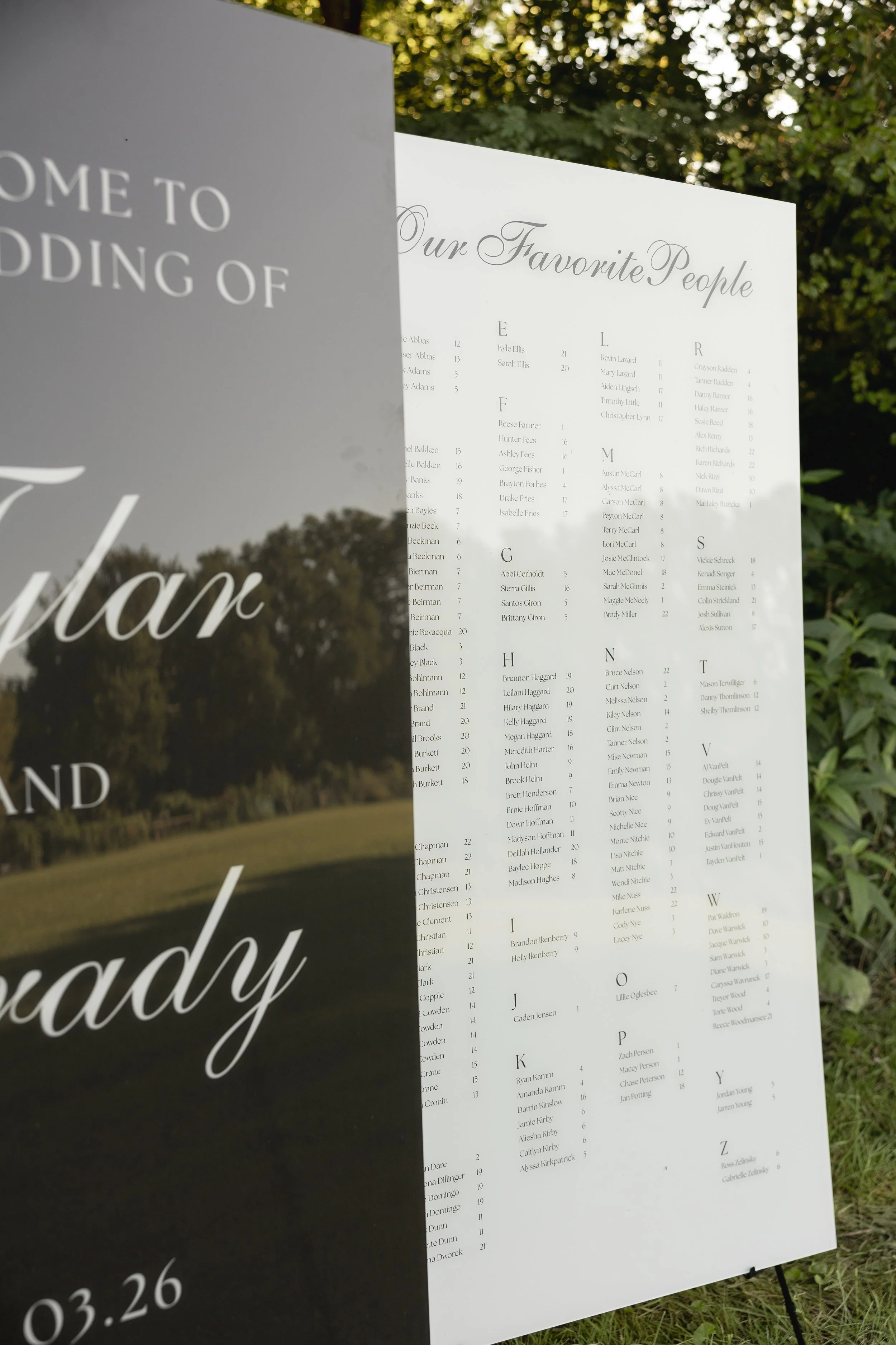An outdoor event sign with a list of names organized alphabetically, featuring a stylish script title 'Our Favorite People' and text indicating a date '03.26' on a black background behind a white panel, with some reflections and trees in the backgrou