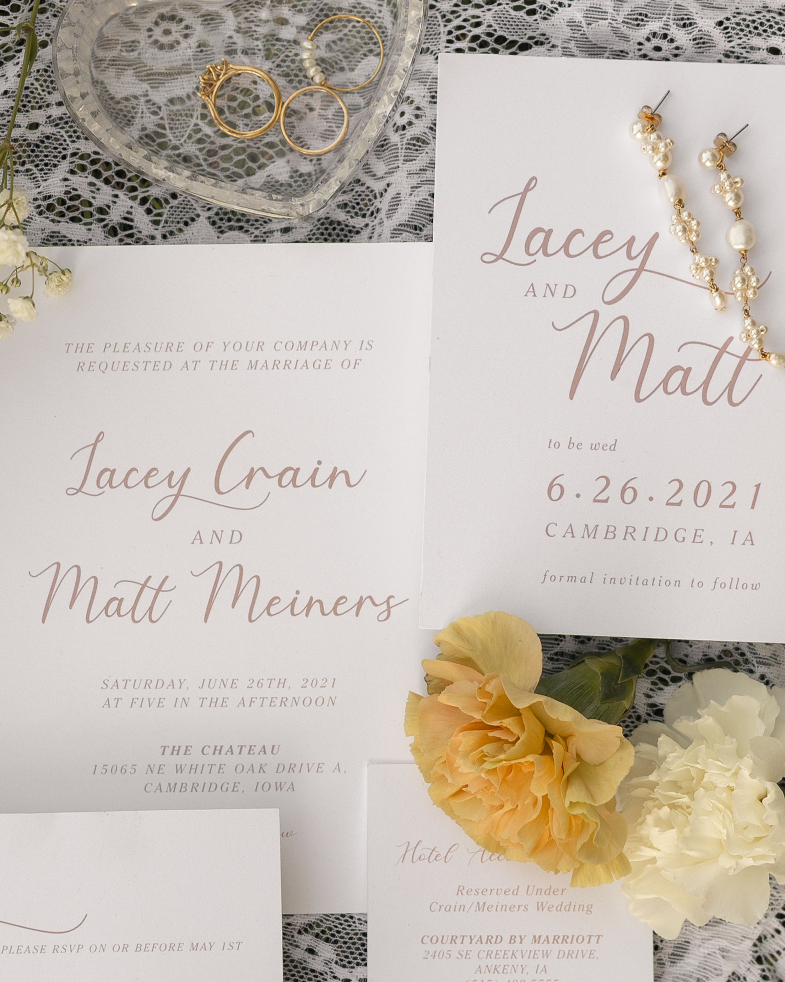 Wedding invitation cards, a pair of pearl and gold earrings, a set of gold rings with pearls, a heart-shaped glass dish, lace fabric, and yellow flowers on a lace tablecloth.