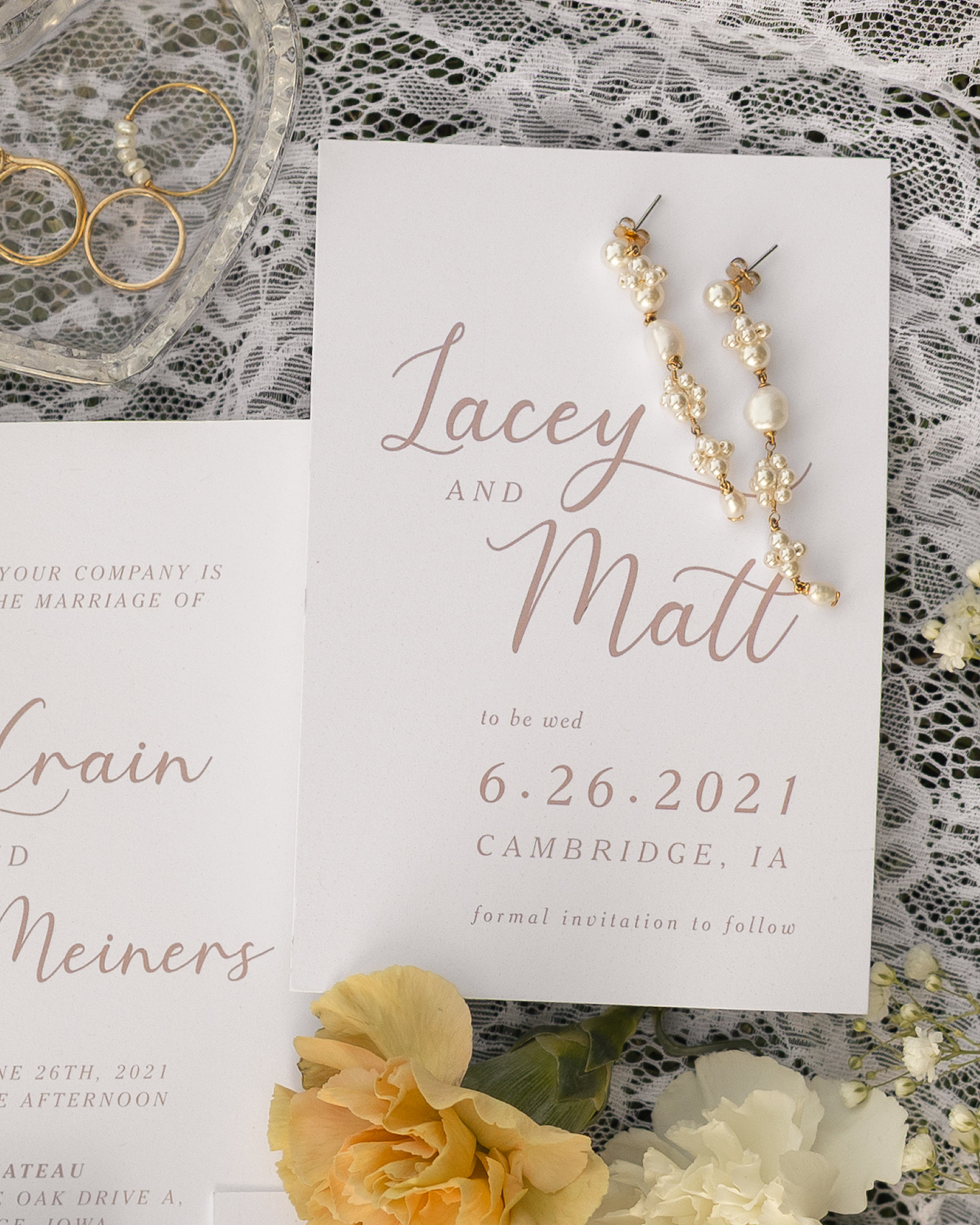 Wedding invitation card with a floral background, pearl and gold jewelry, and a card with the names "Lacey and Matt" along with wedding details including date, location, and calligraphy text.