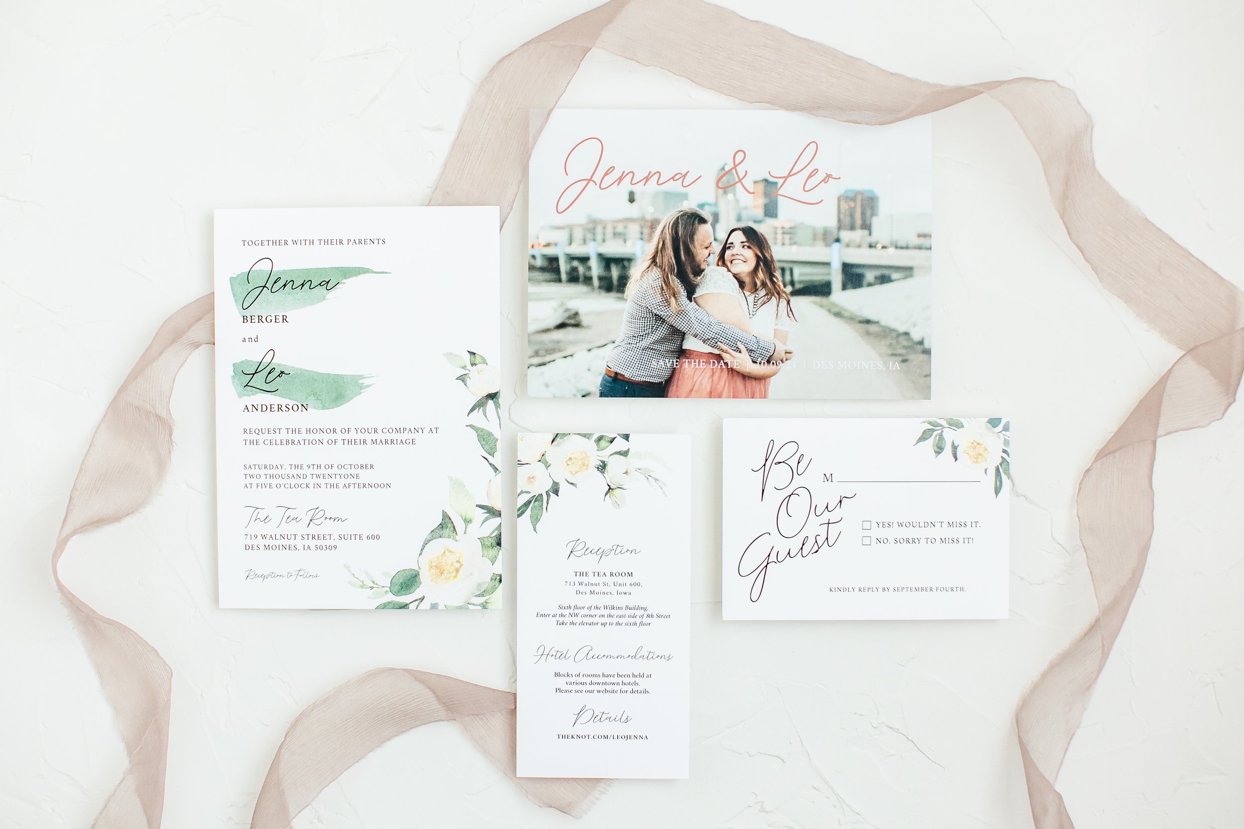 Wedding invitations and RSVP cards on a white surface, surrounded by beige ribbon, featuring a photo of a smiling couple hugging on a bridge with city skyline in the background.