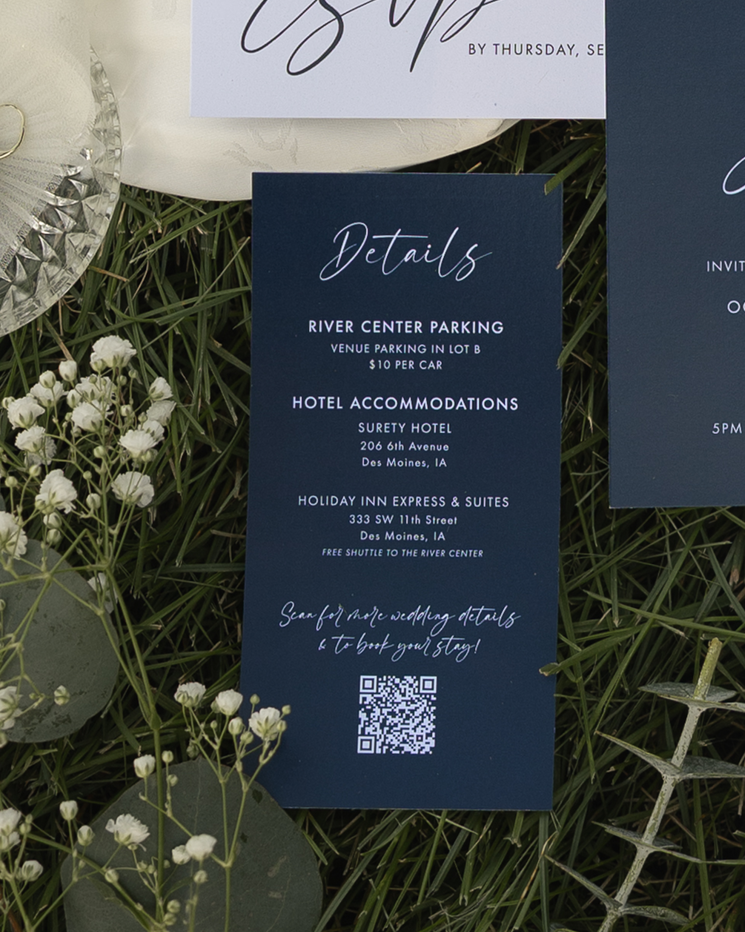 A wedding invitation with details about parking and accommodations, placed on grass with small white flowers nearby. The invitation has a dark blue background with white and elegant script text, and a QR code at the bottom.
