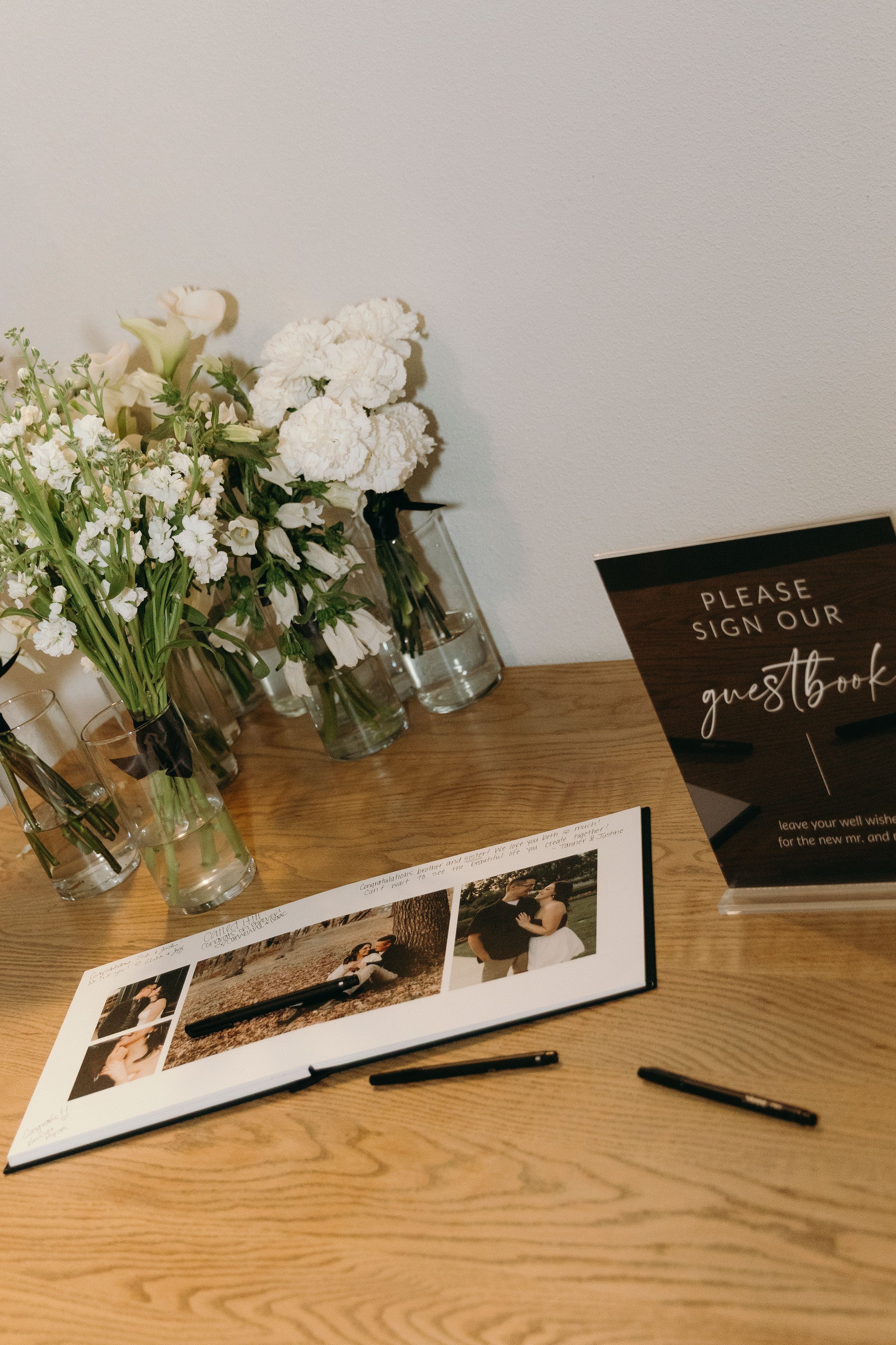 A table set up with various flower arrangements in glass vases, a guest book with photographs and pens, and a sign that says "Please sign our guest book." The flowers include white roses and other white blooms, and the background is a plain wall.