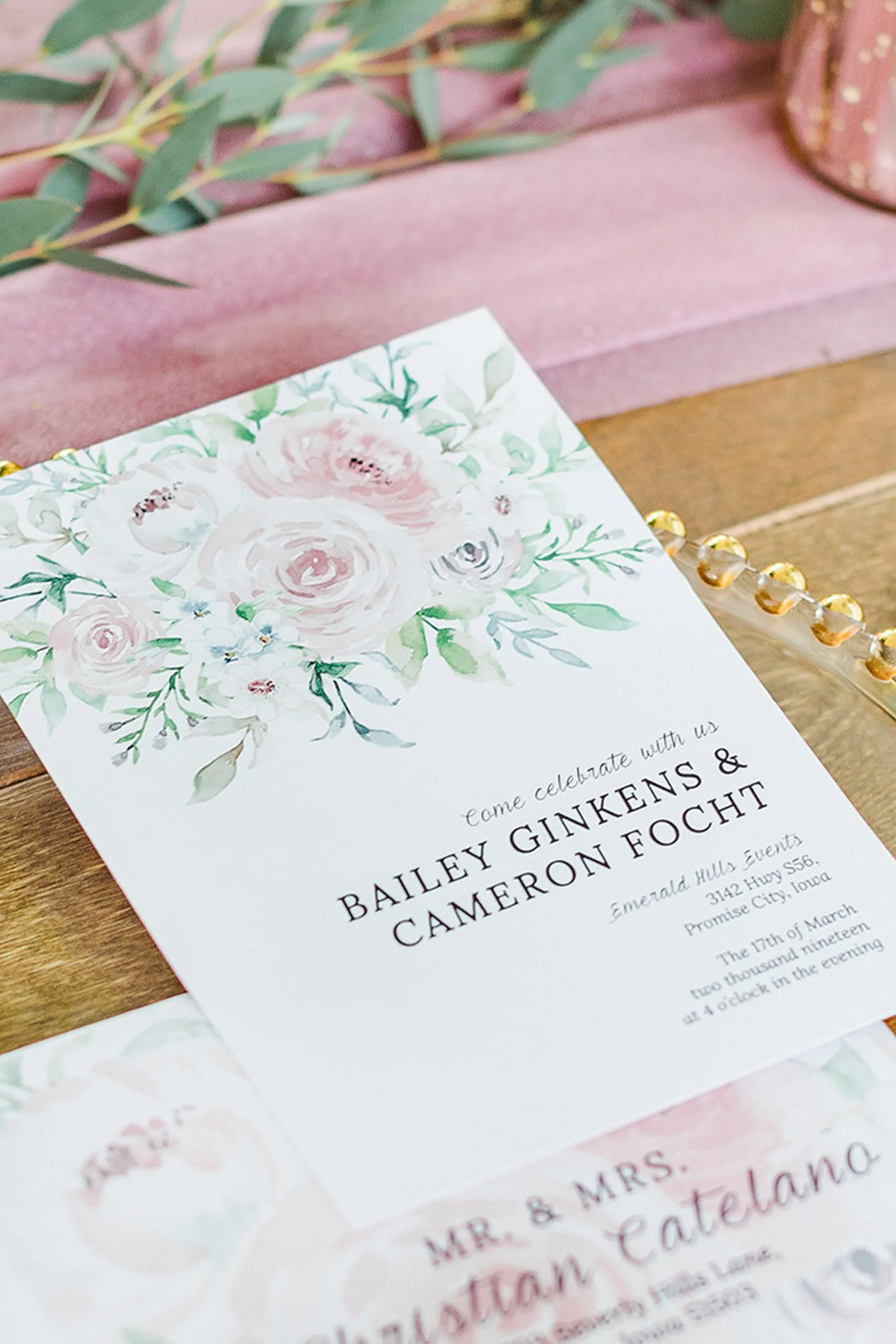 A floral wedding invitation with soft pink and green watercolor flowers at the top. The invitation invites guests to celebrate the wedding of Bailey Ginkens and Cameron Focht at Emerald Hills Events in Iowa, scheduled for March 17th at 4 o'clock in t