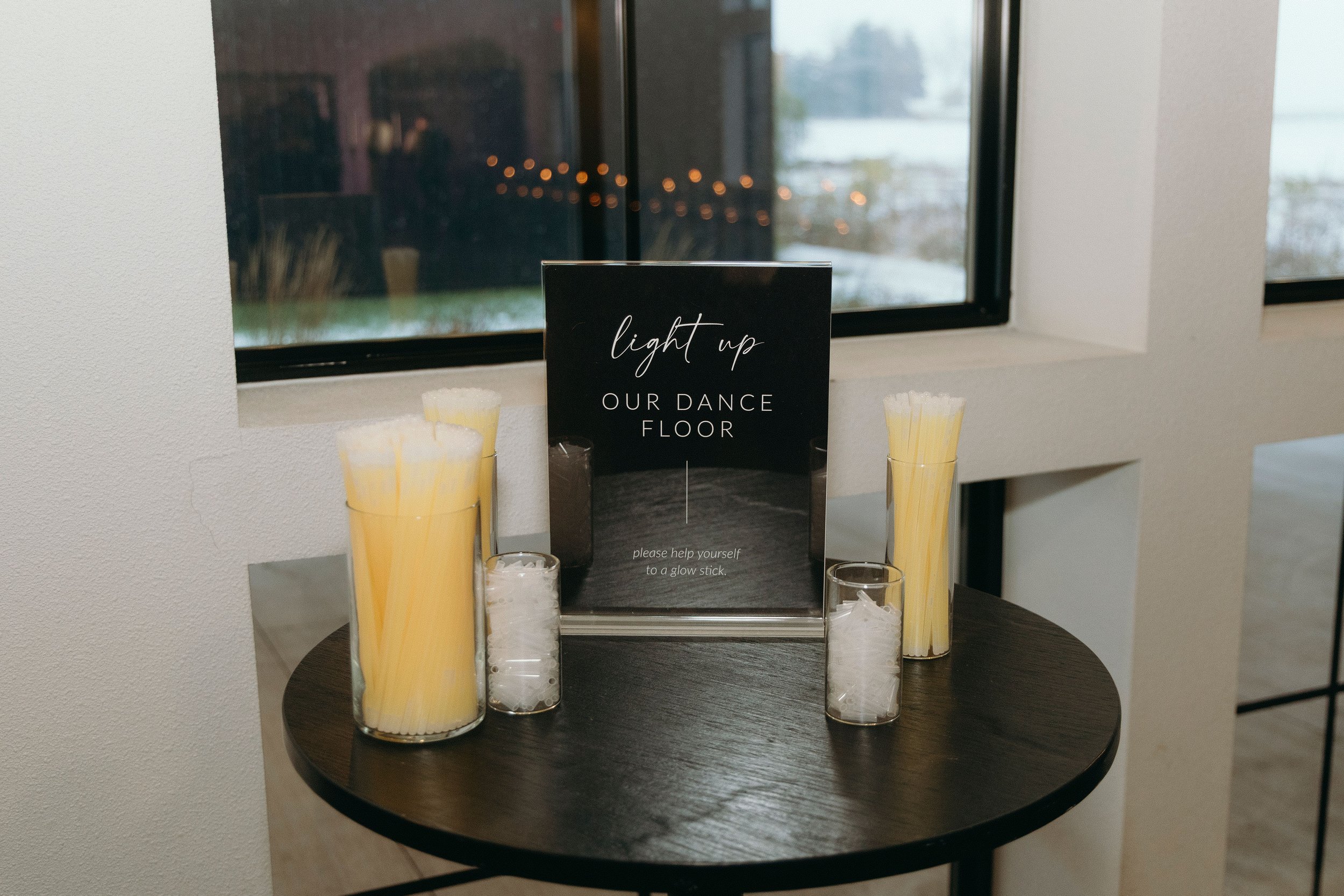 A round black table with multiple white candles in glass containers and tall yellow candles in taller glass holders. A black sign with white cursive text that says 'light up OUR DANCE FLOOR' and smaller text underneath reading 'please help yourself t