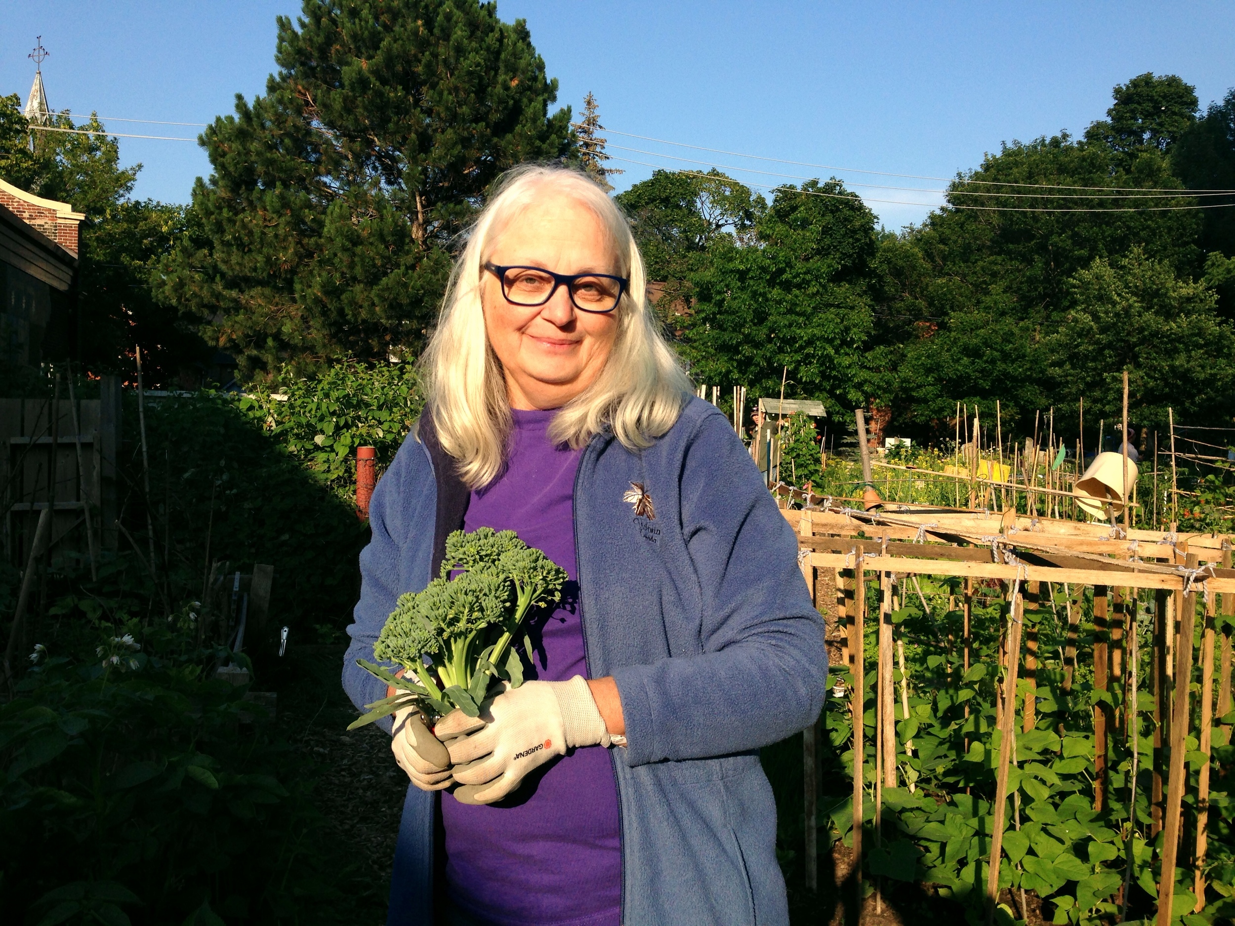  A woman standing in front of HOPE Community Garden Plots, holding a crown of broccoli and smiling 