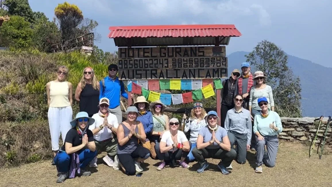 A smiling group of yoga retreat participants posing together by the Hotel Gurans Australian Camp sign during a mountain trekking excursion.