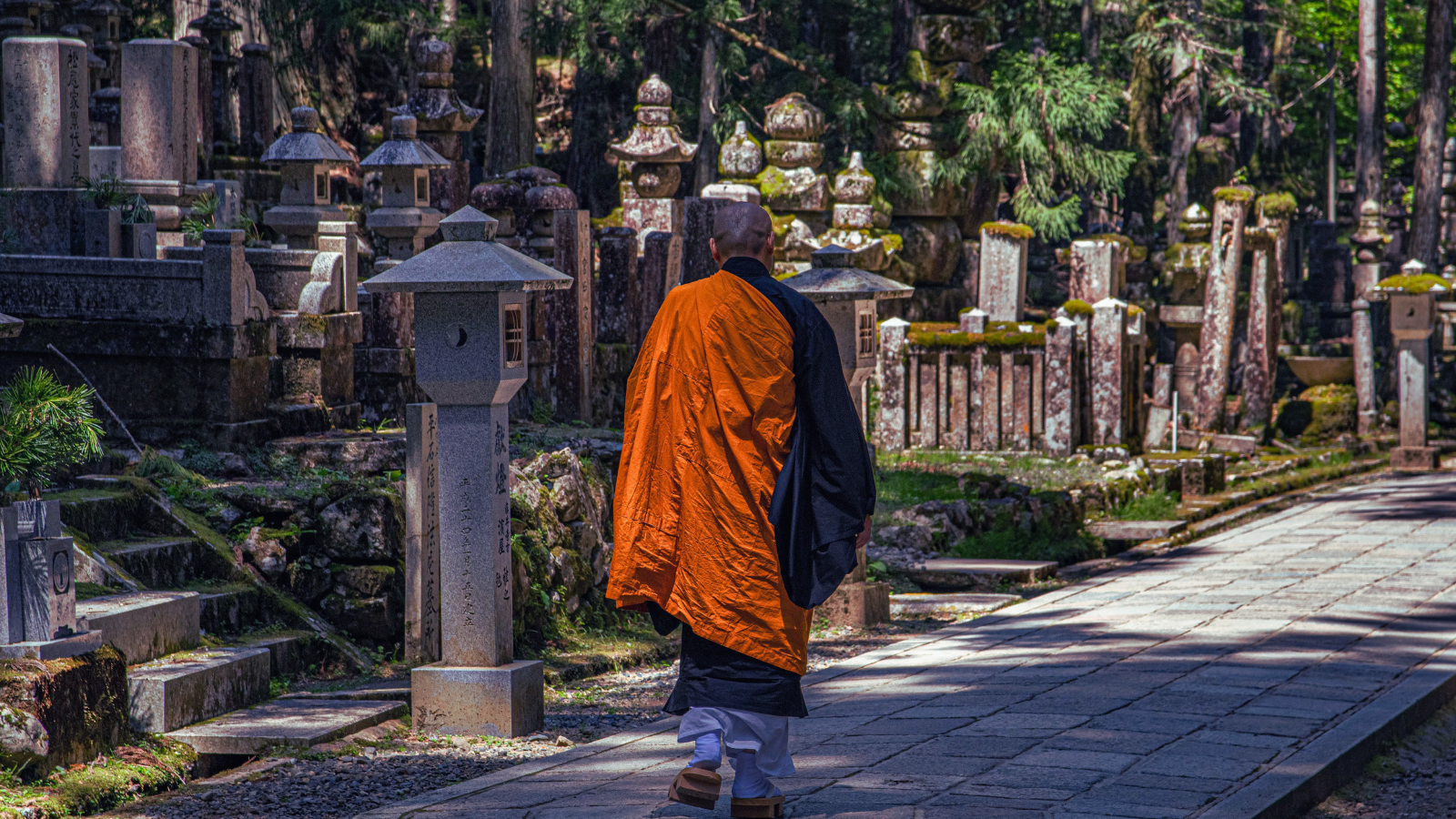 A Buddhist monk in a vibrant orange robe walking along a stone pathway lined with lanterns and mossy stupas in Koyasan, Japan.