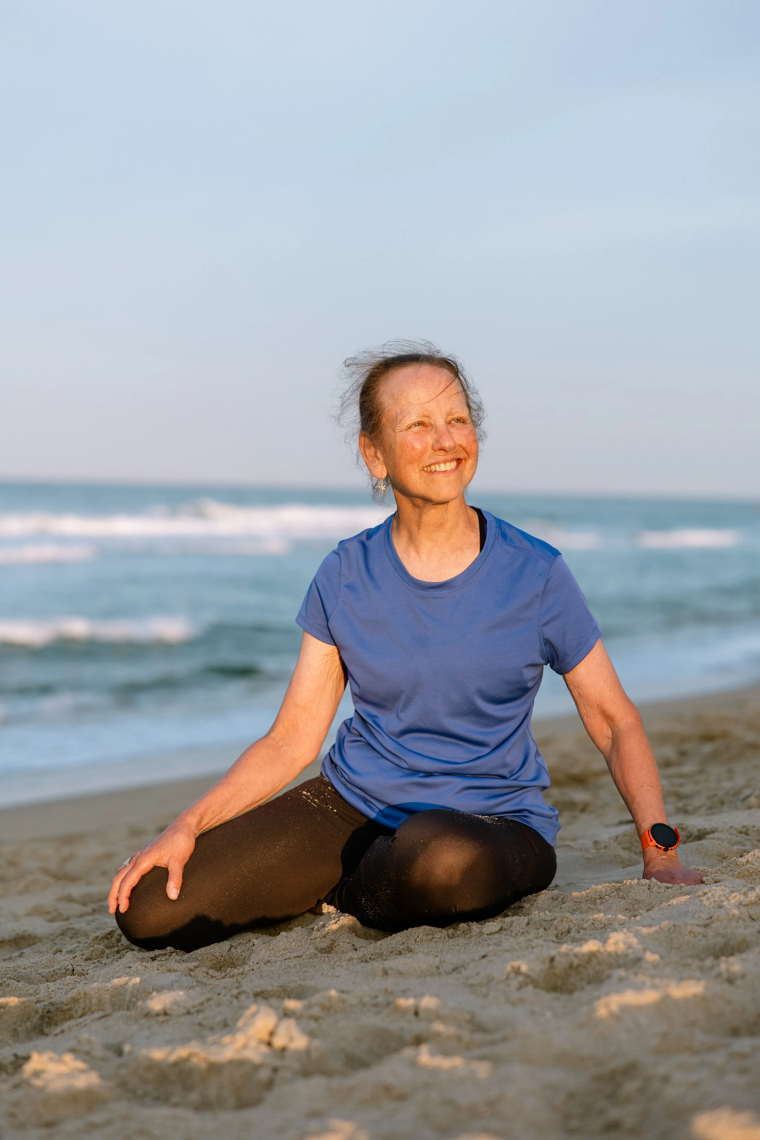 Outer Banks Yoga instructor Kathy Cagiano seated in a yoga pose on the sandy beach at Kitty Hawk with ocean waves in the background.