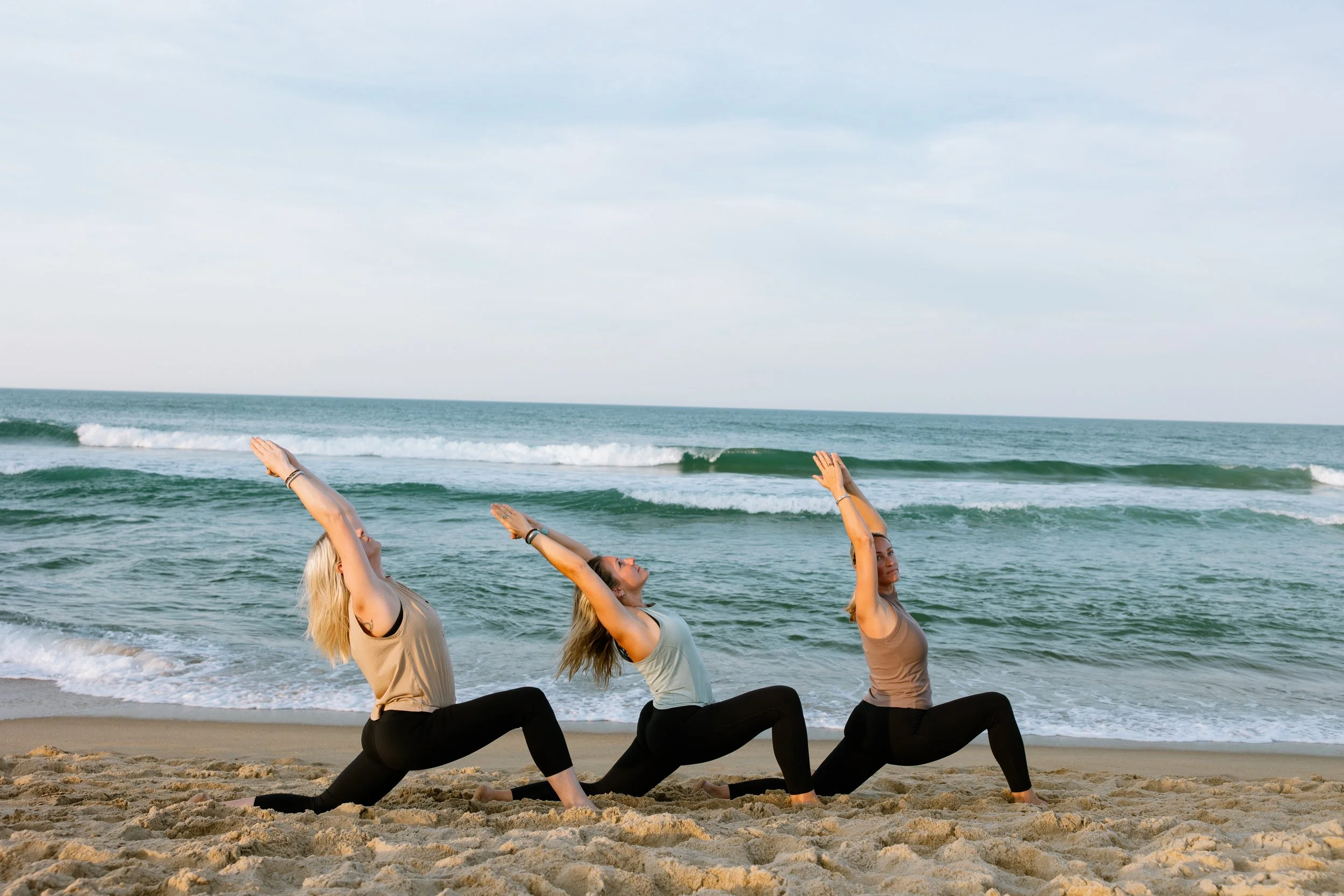 Three women in low lunge pose at the shoreline on the beach near the Kitty Hawk Pier in Kitty Hawk NC