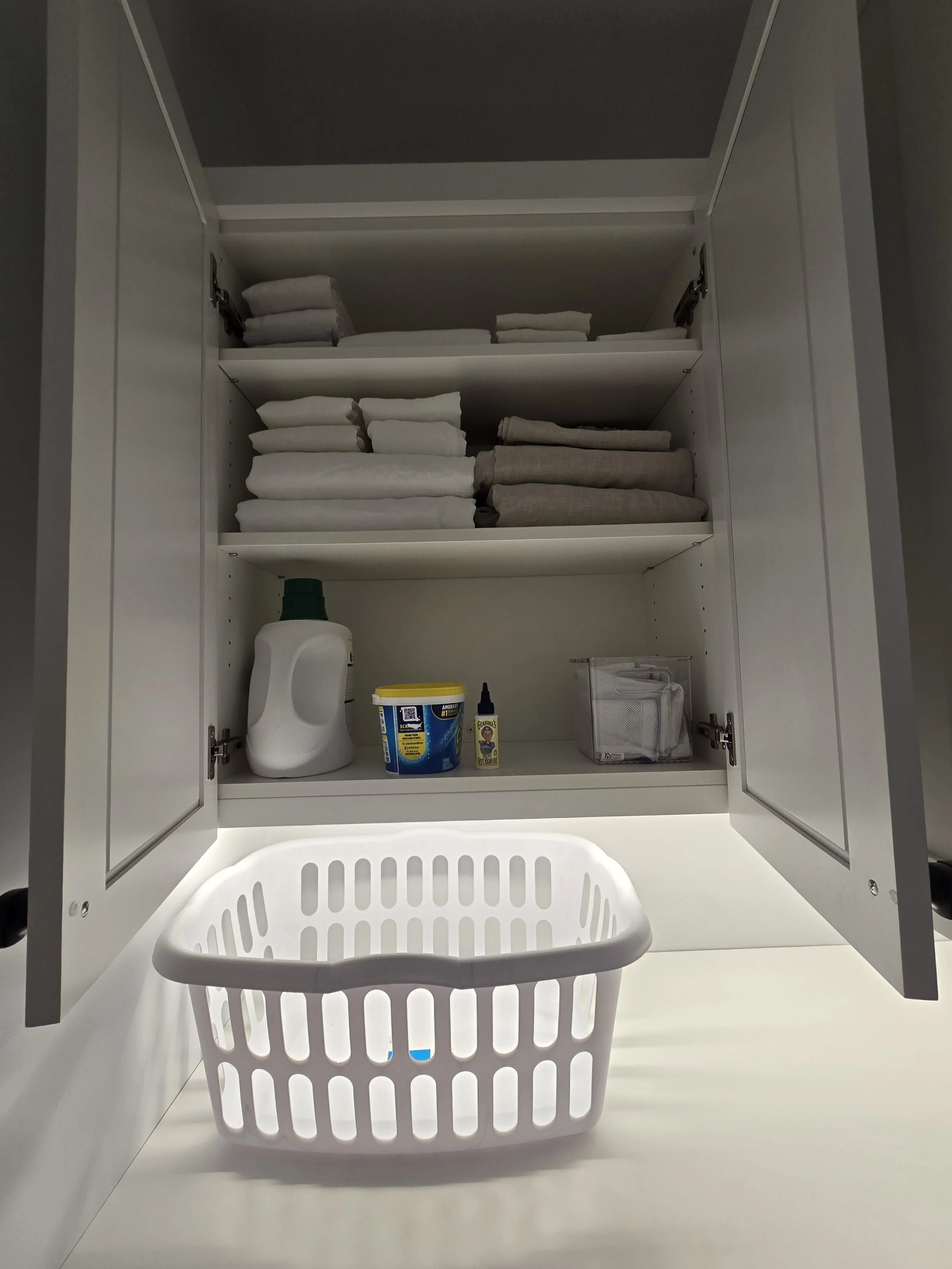 An open white kitchen cabinet with folded towels and cleaning supplies inside, and a white laundry basket on the countertop.