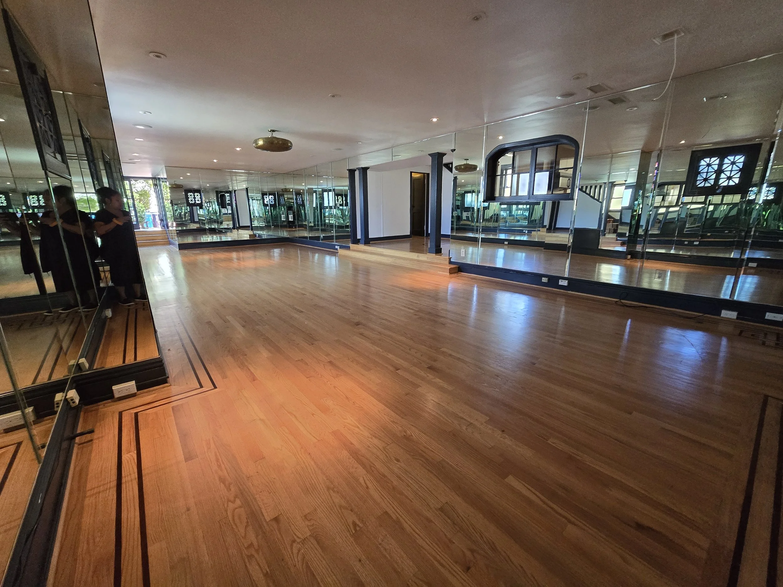Empty dance studio with hardwood floor and mirrored walls, two children standing in the corner, sunlight streaming through windows.