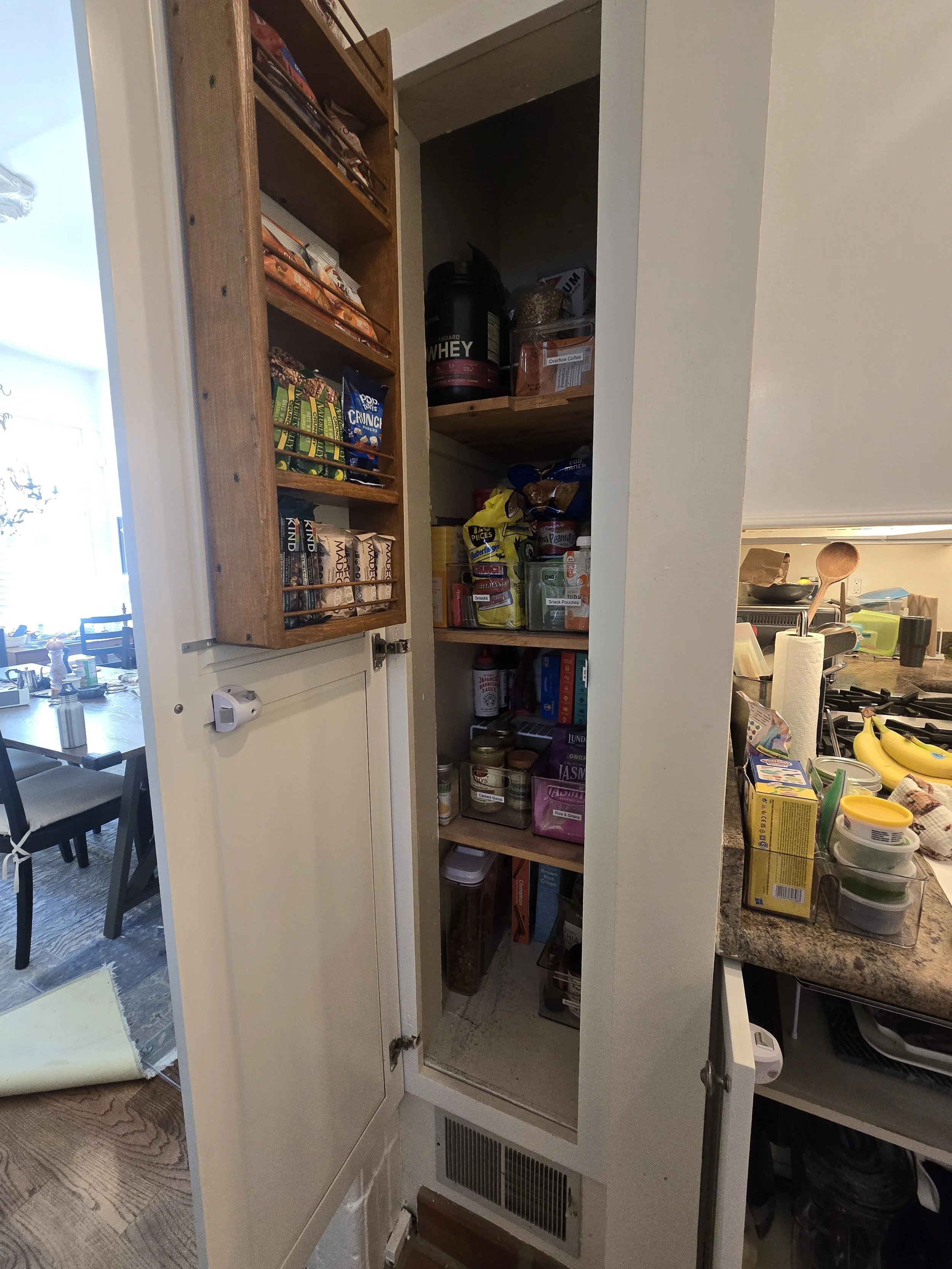 Open pantry closet with various snacks, cereals, canned goods, and dietary supplements. Adjacent to kitchen with bananas, containers, and paper towel roll on countertop.