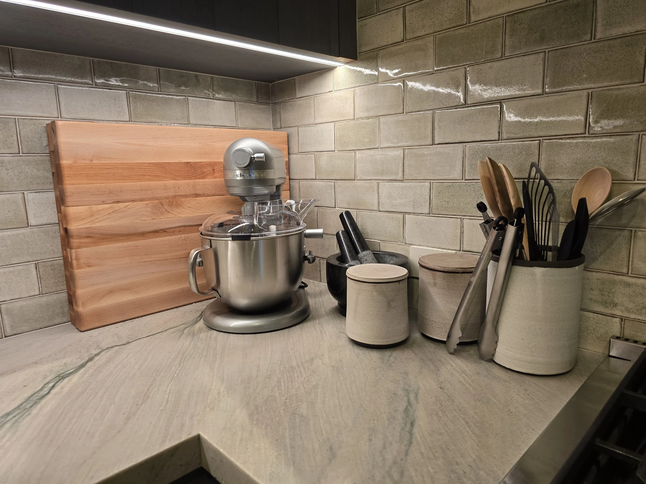 Kitchen countertop with a stand mixer, a wooden cutting board, storage jars, a mortar and pestle, and a utensil holder with wooden and black utensils against a brick backsplash.