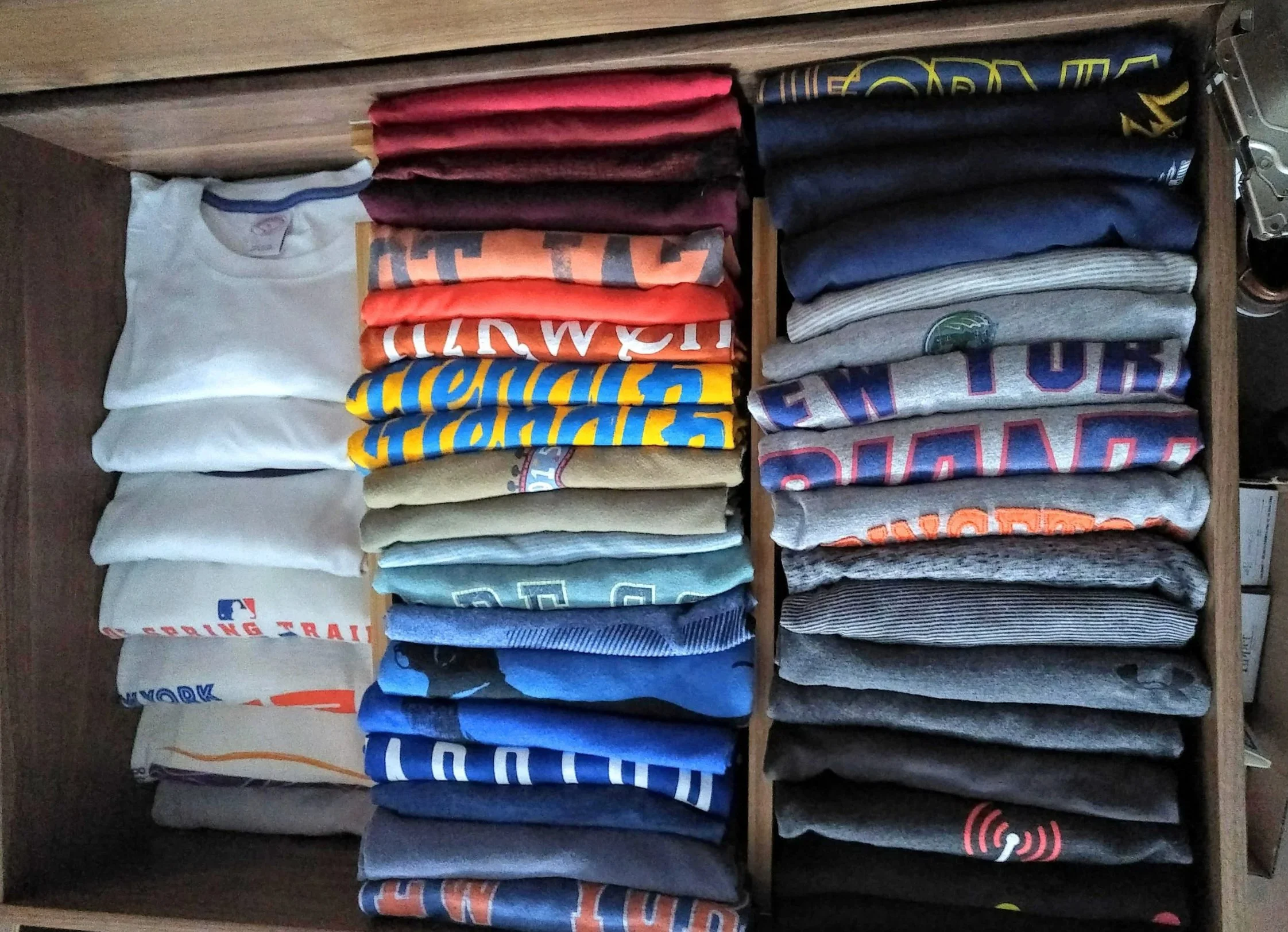 Stacks of folded T-shirts in various colors and designs inside a wooden drawer.