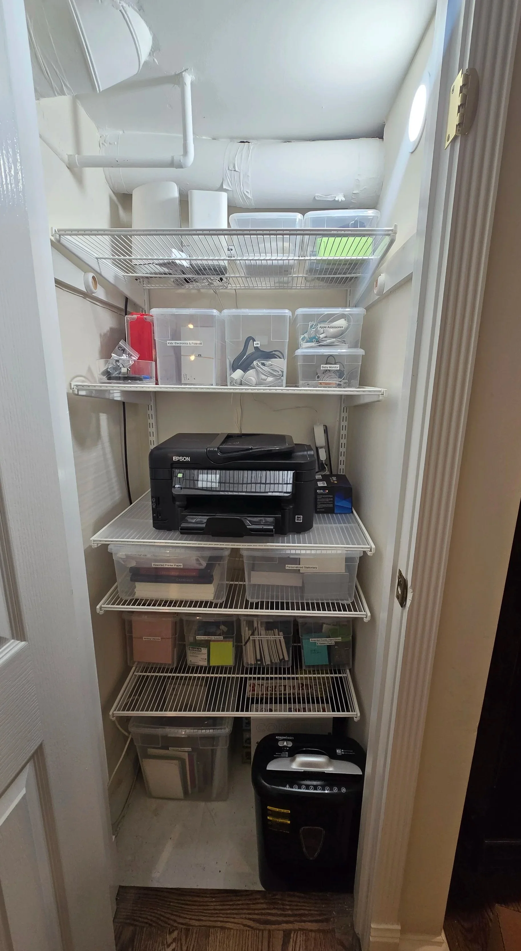 A small walk-in storage closet with shelves holding various containers, a printer, and office supplies.