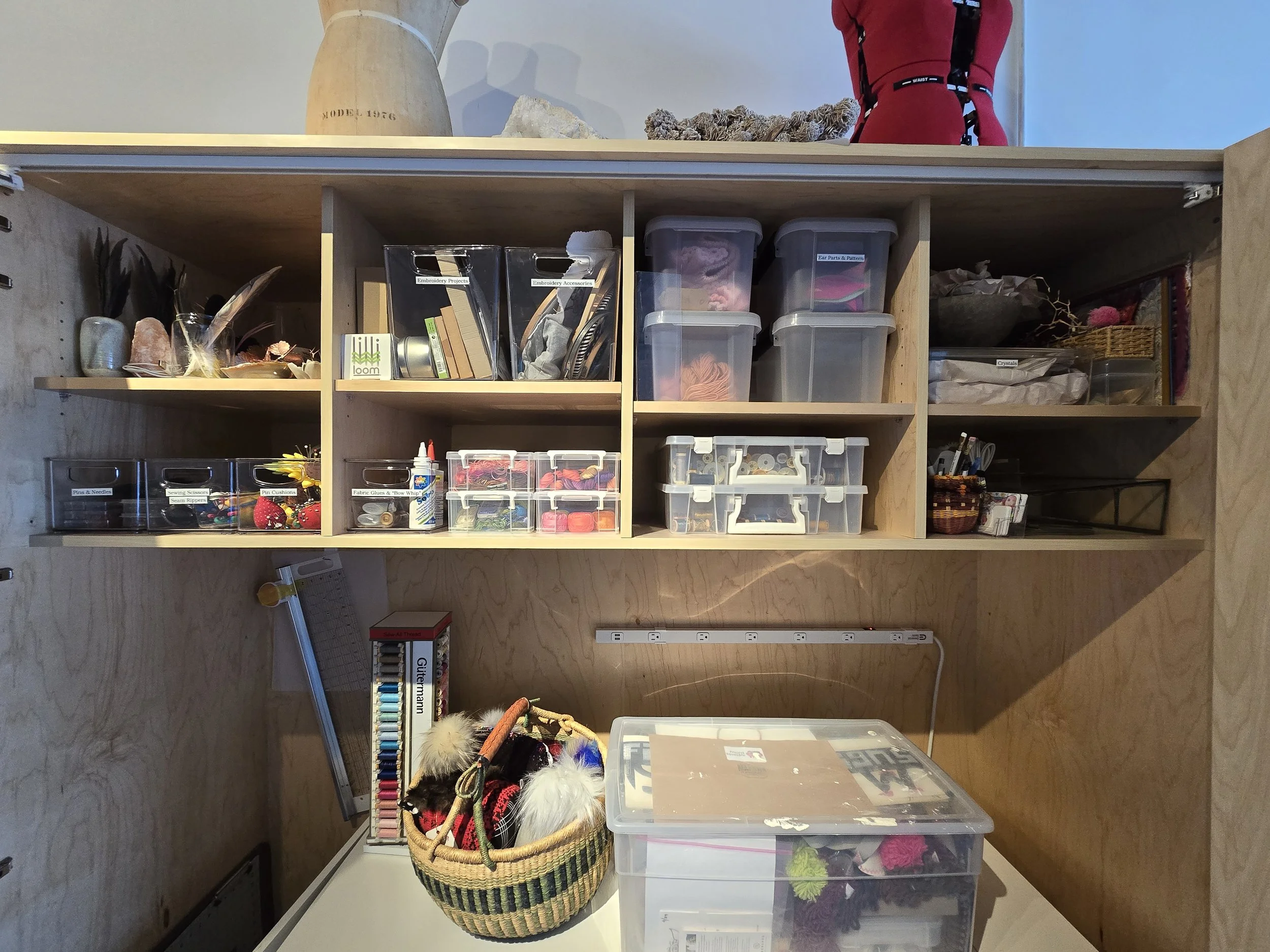A well-organized craft storage area with wooden shelves filled with various craft supplies, including containers of yarn, buttons, fabric glue, and other sewing and crafting tools. A basket with felted animals and a box of thread are on a table in fr