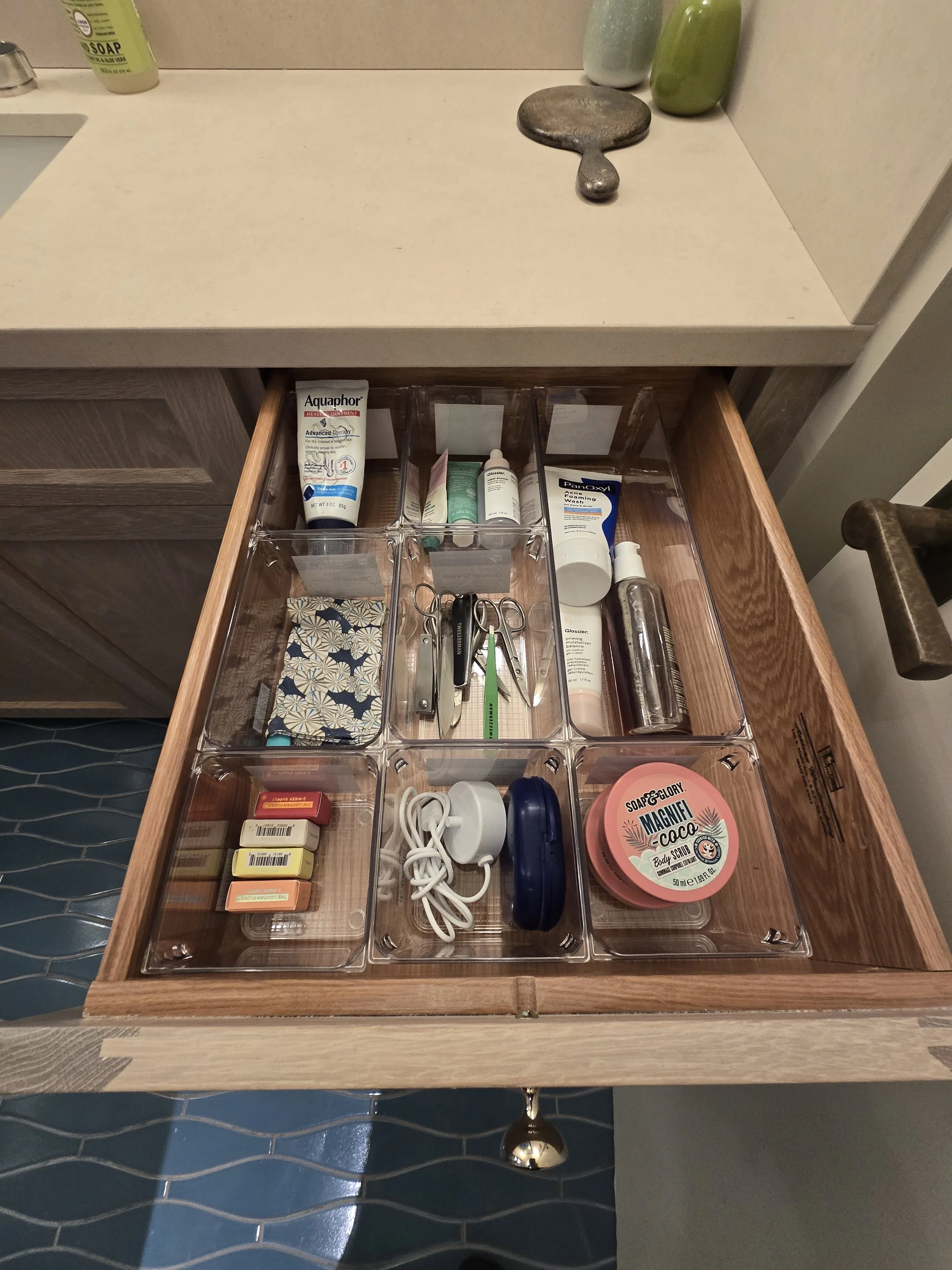Open drawer with acrylic compartments containing toiletries and personal items, including lotion, scissors, chapstick, hair ties, a jar of body scrub, and other small personal care products.