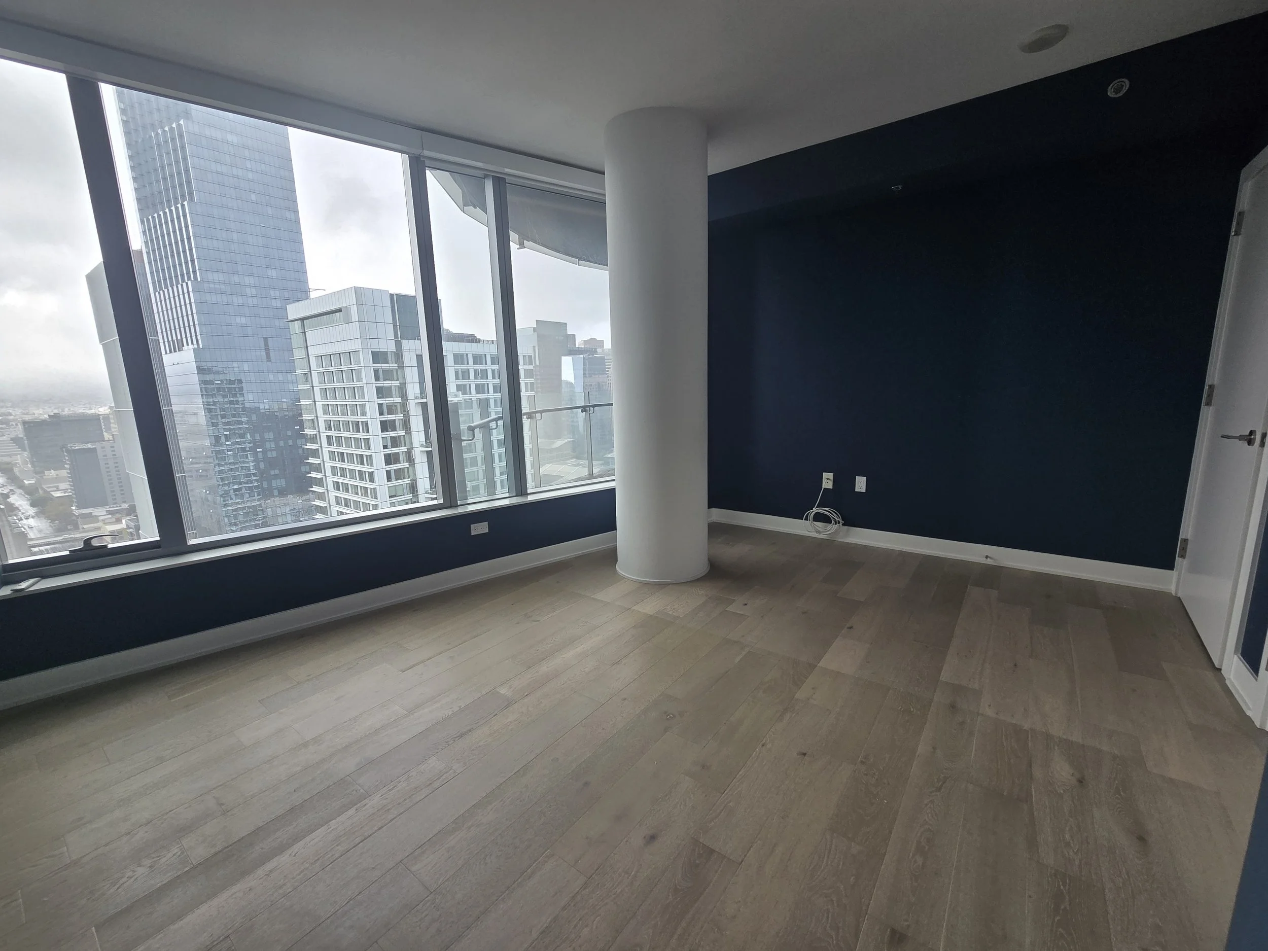 Empty modern high-rise apartment room with large windows overlooking city skyline; dark blue accent wall, light wood flooring, and white door.
