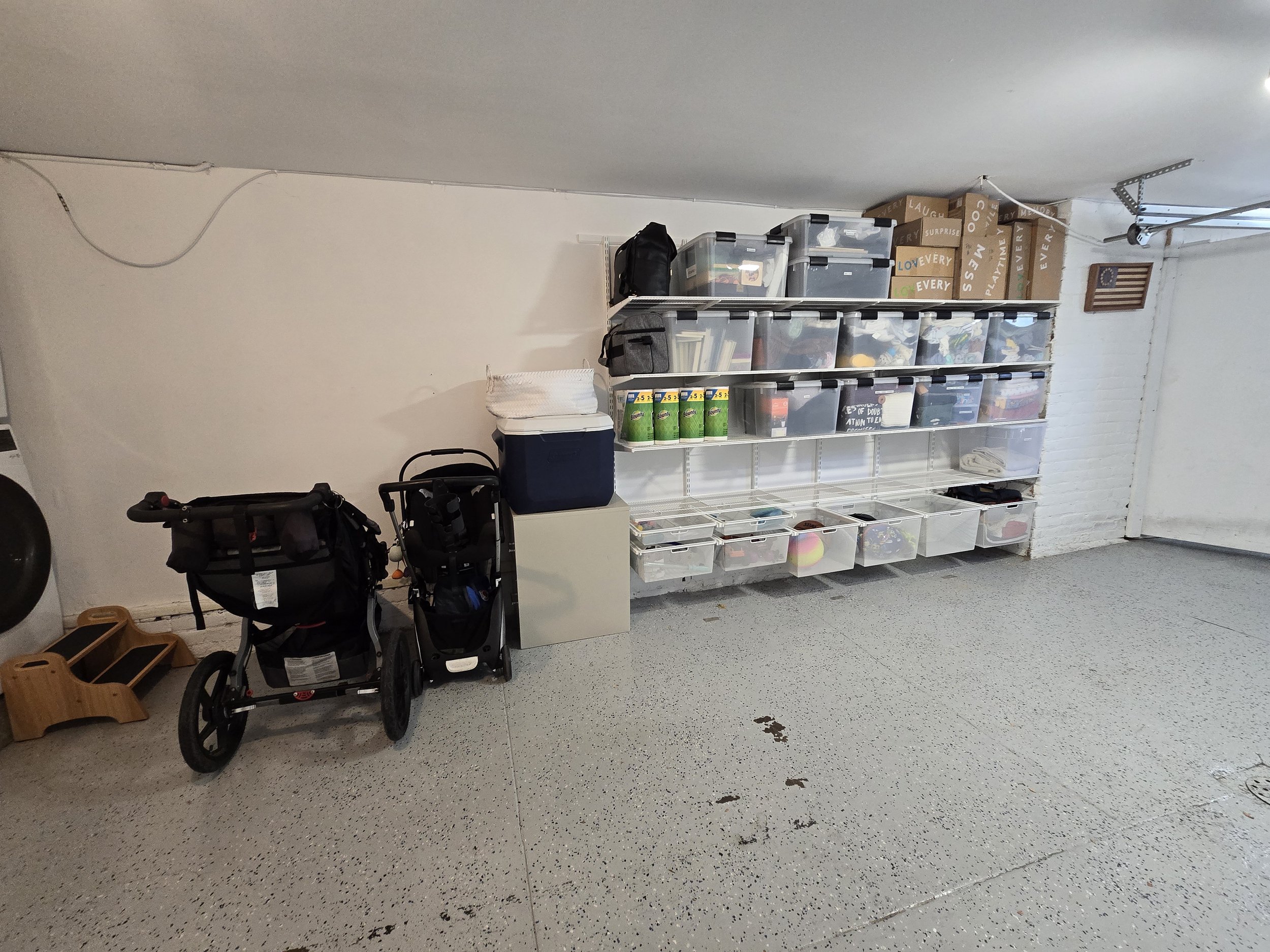 A garage storage area with clear plastic bins on white shelving, two black strollers, a small white cabinet, and some boxes on the shelf, with a speckled gray floor and white brick wall.