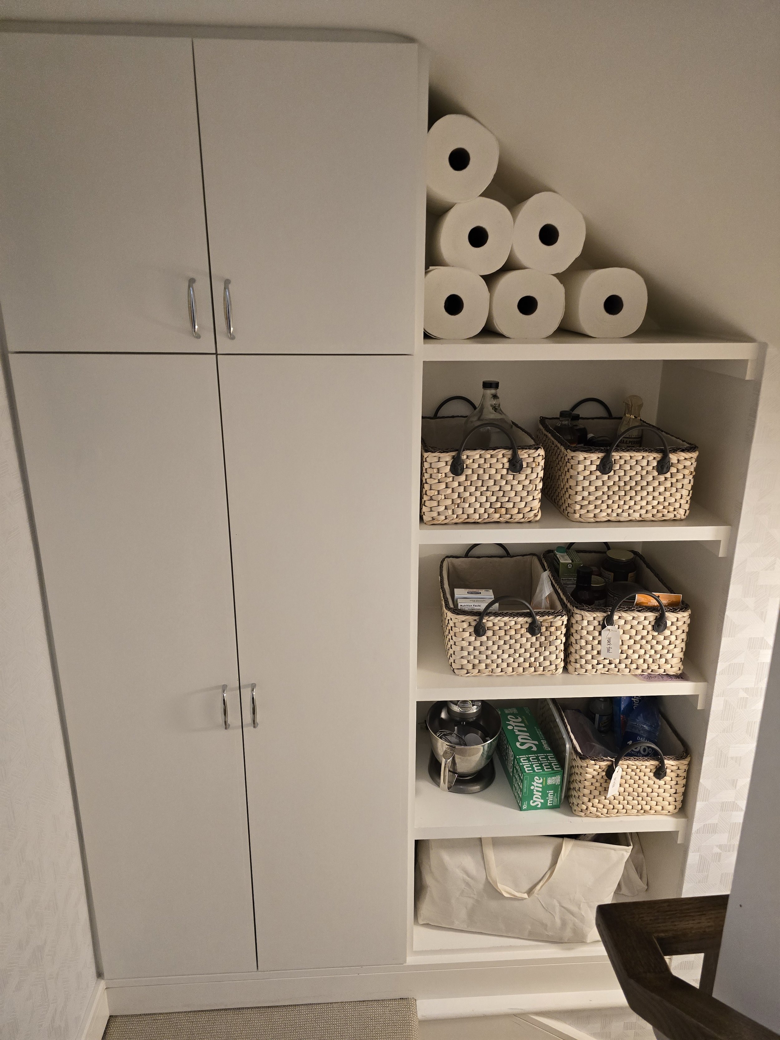 White storage cabinet and open shelving unit with paper towels, woven baskets, and miscellaneous household items in a kitchen.