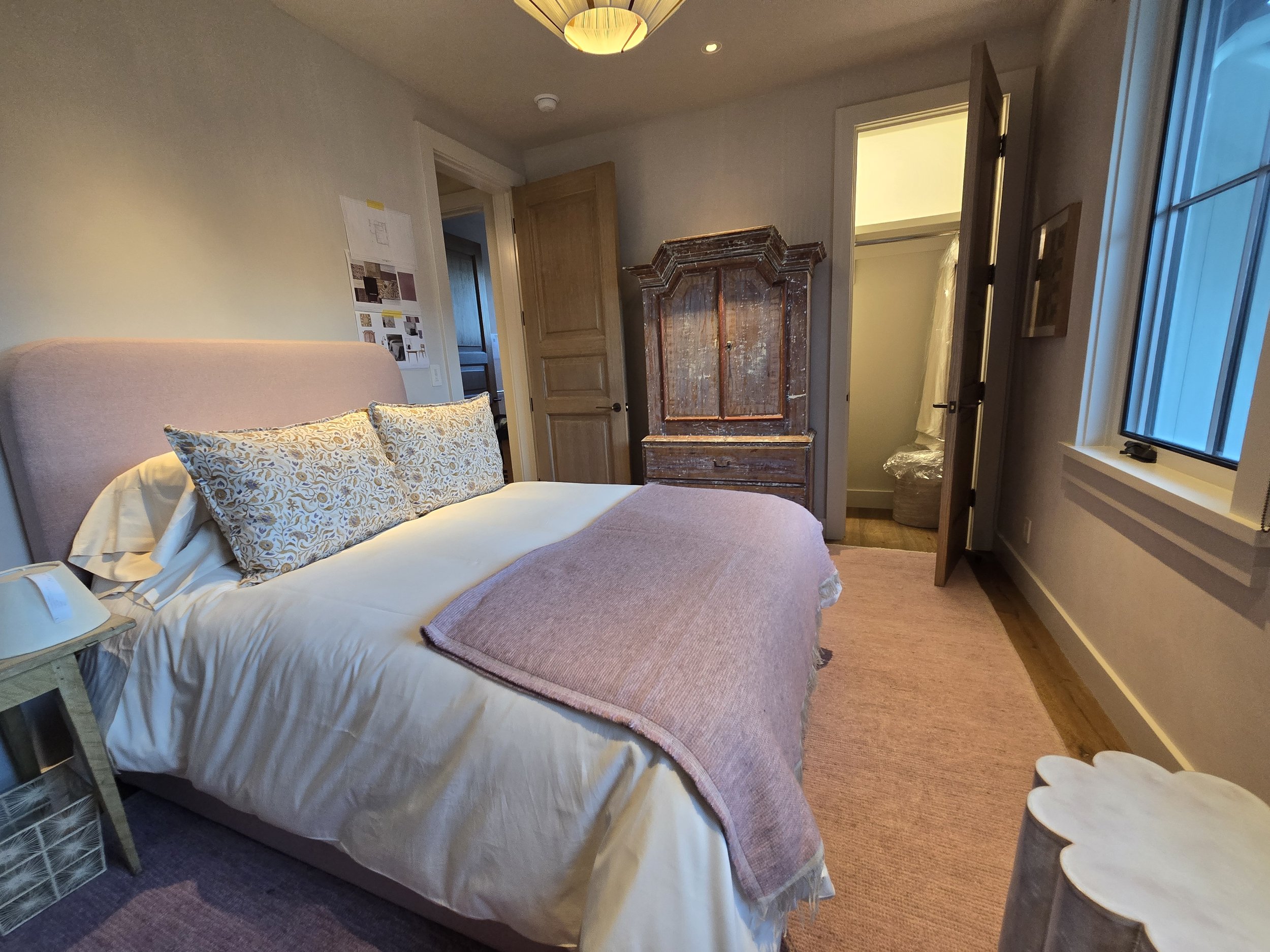 A cozy bedroom with a pink upholstered headboard, a bed with white and patterned pillows, a pink throw blanket, a wooden nightstand with a lamp, an antique wardrobe, an open door revealing a laundry area, a window with blue hue, a pink area rug, and 