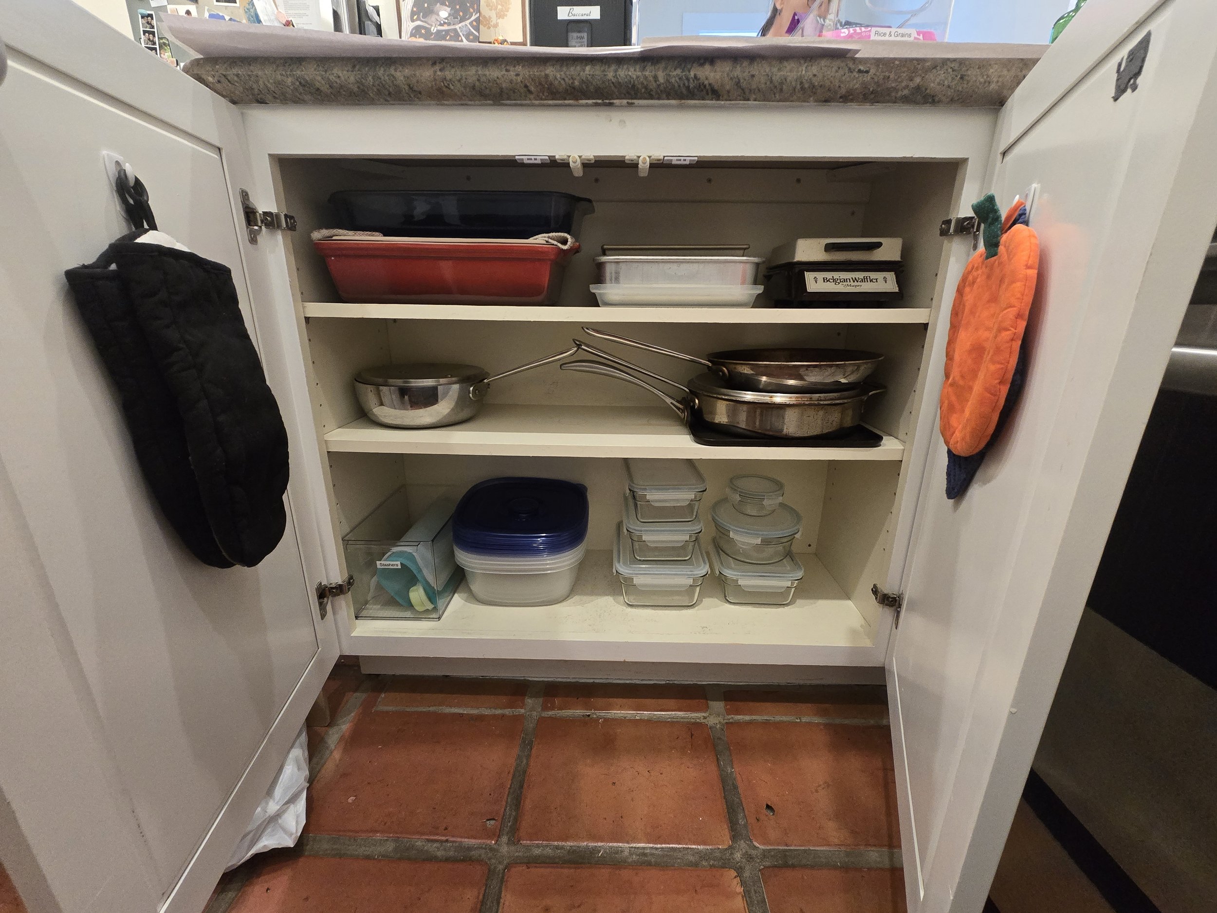 Open kitchen cabinet with various pots, pans, storage containers, and a mitten hanging on the door