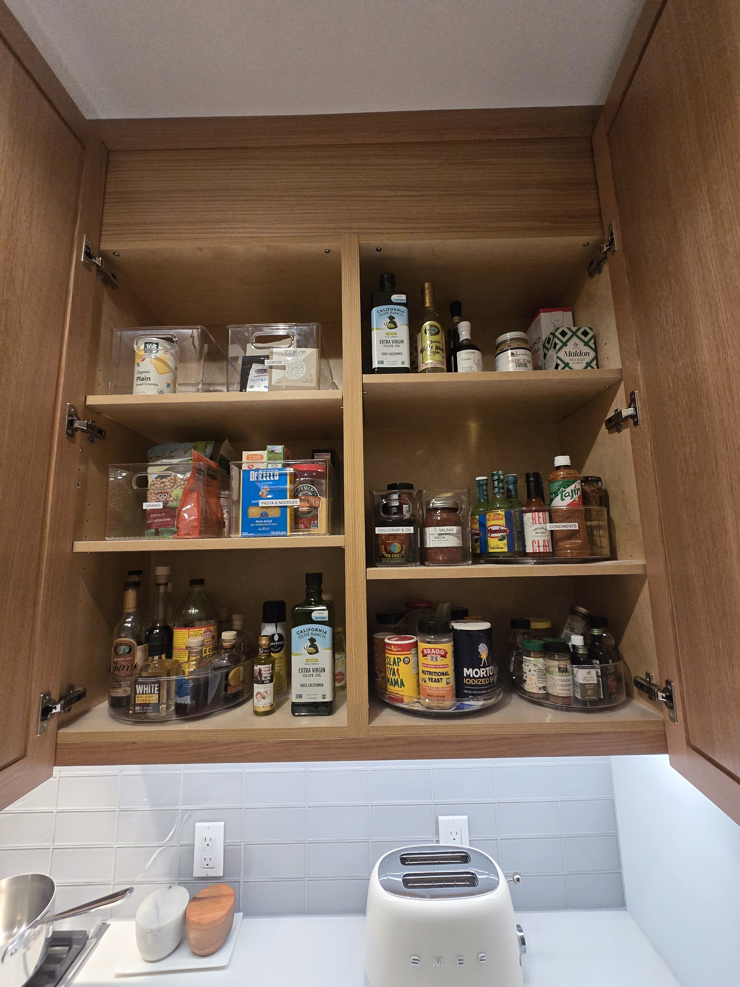 Open kitchen cabinet with various bottles, jars, and containers of spices, herbs, and cooking ingredients inside, above a white countertop with a toaster and some mugs.
