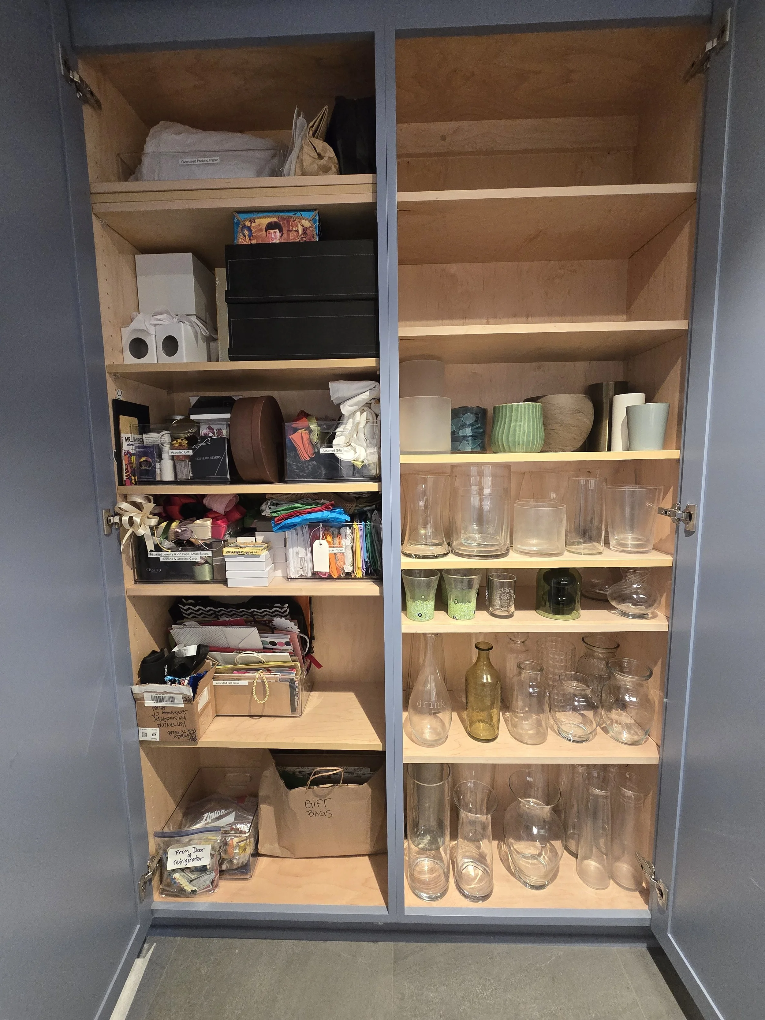 A storage cabinet with shelves, containing various items like glassware, boxes, and miscellaneous household objects.