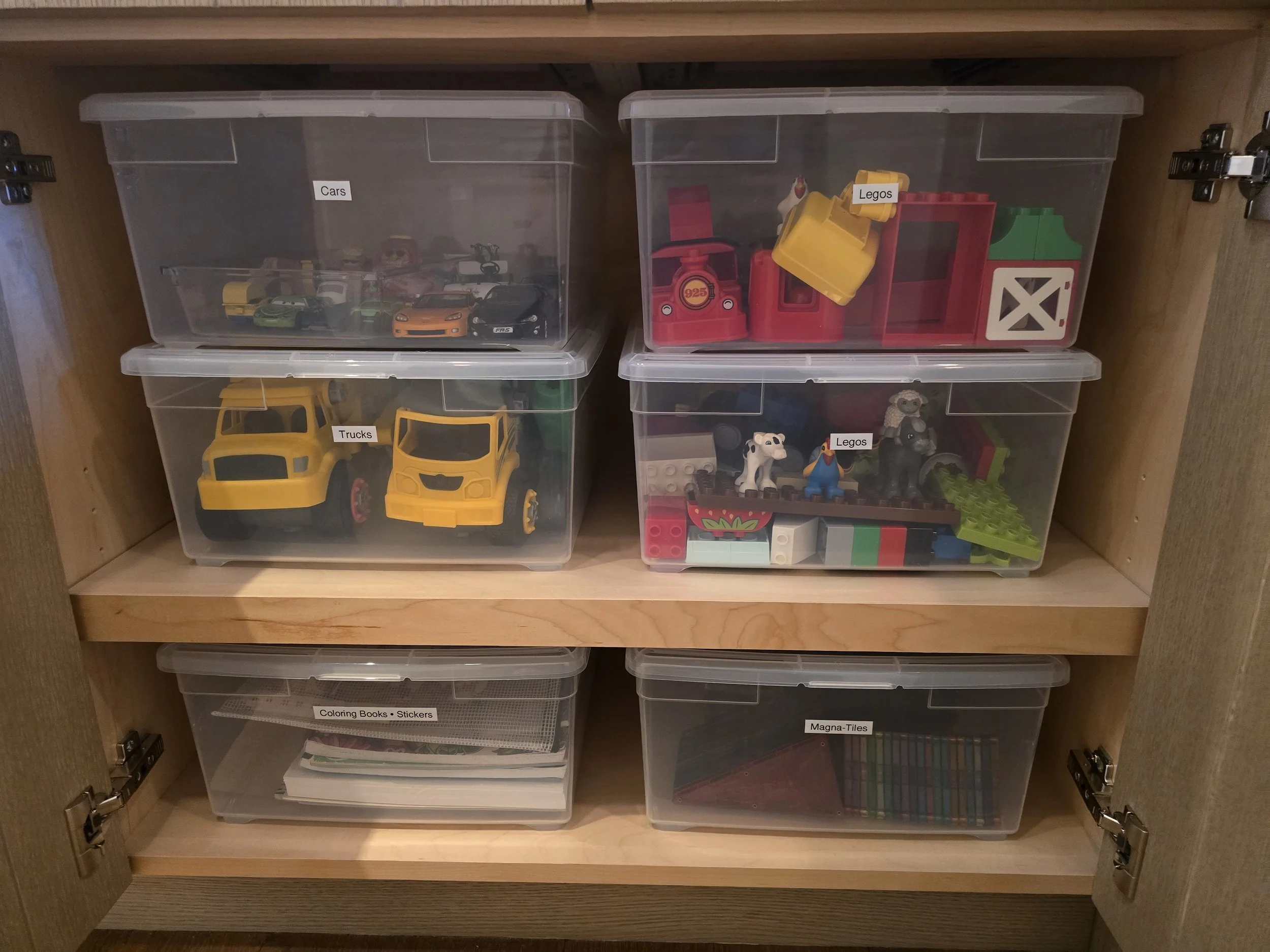 A wooden cabinet with four plastic bins labeled 'Cars,' 'Trucks,' 'Legos,' 'Coloring Books / Stickers,' and 'Magna-Tiles,' containing toy cars, trucks, Legos, and coloring materials.
