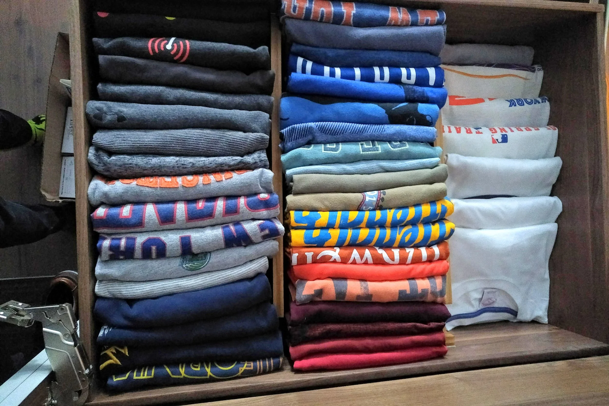 Stacks of folded T-shirts in various colors and designs inside a wooden drawer.