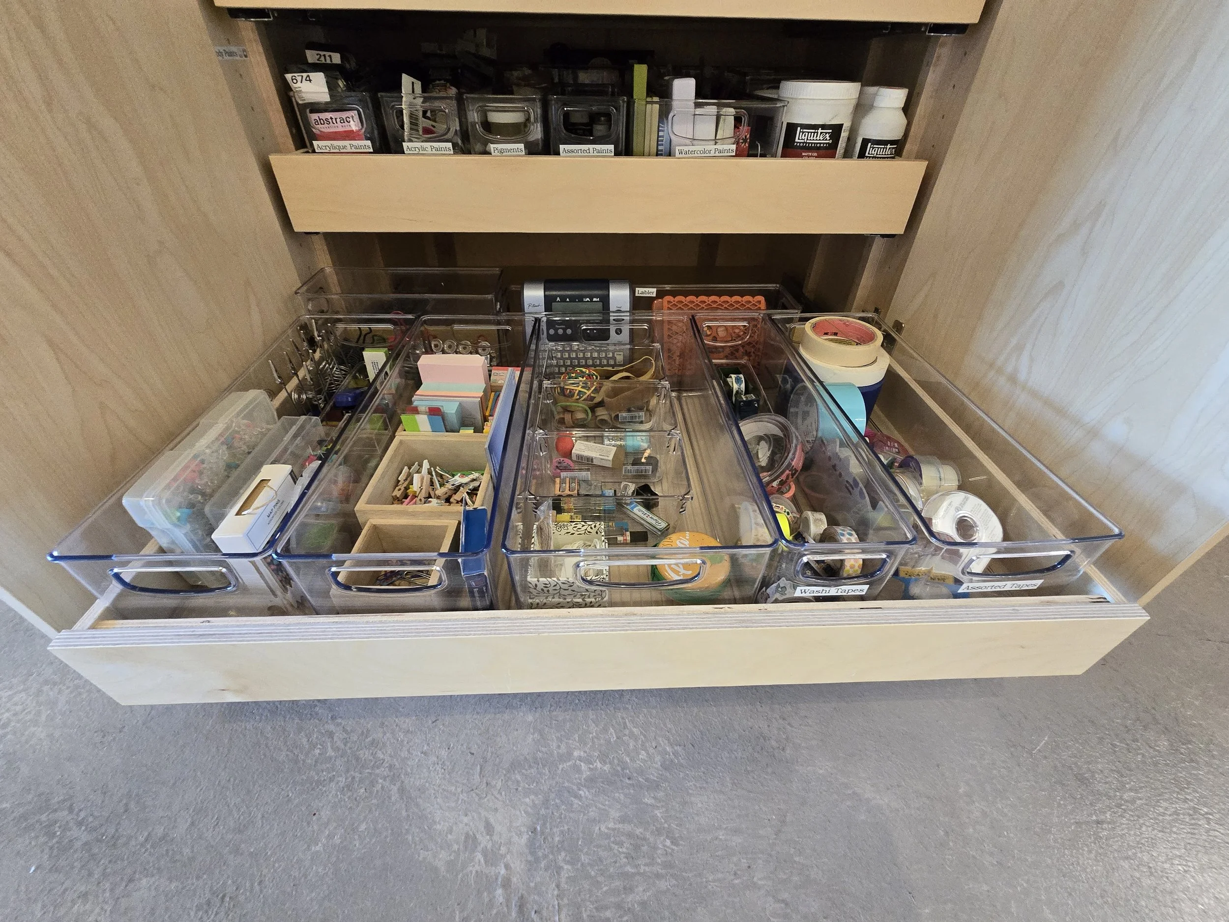 Open drawer with organized clear plastic bins filled with various office supplies such as tape, clips, sticky notes, and cables, with a wooden shelf above holding jars labeled with paint and art supplies.