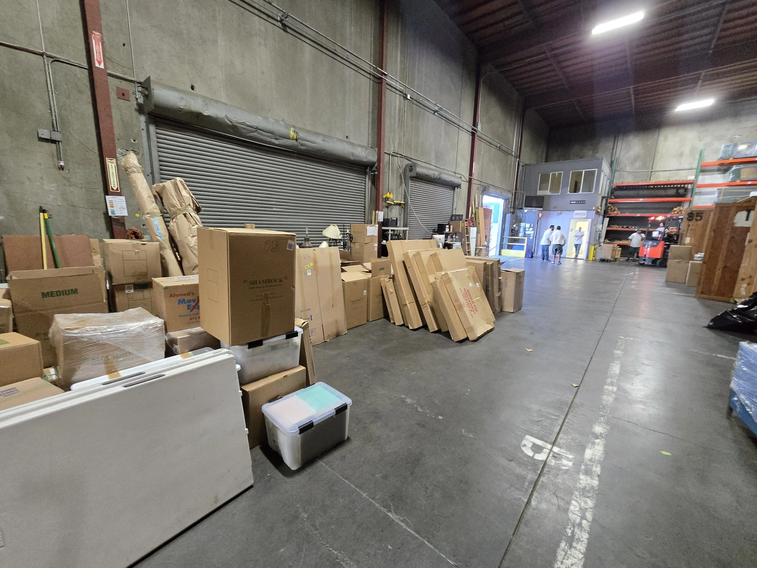 Inside a warehouse with cardboard boxes stacked along one side and a group of people near the back entrance.