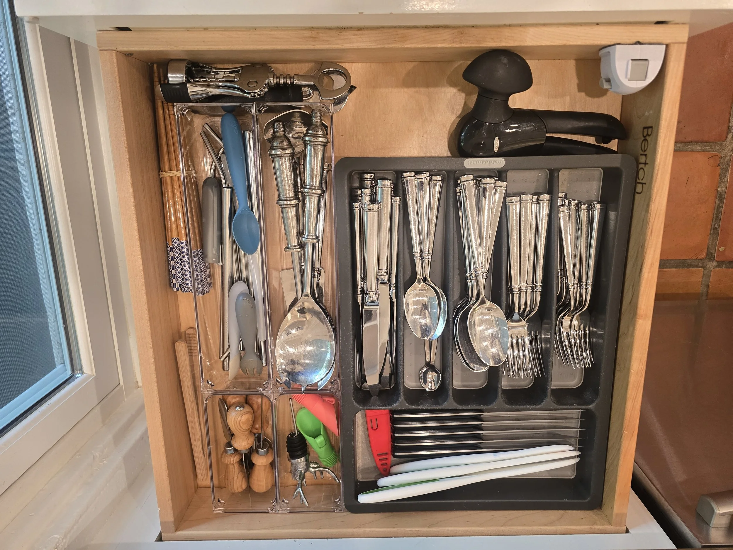 Kitchen drawer containing various utensils, including spoons, forks, knives, vegetable peelers, whisks, a garlic press, and a meat tenderizer. Some utensils are stored in a black utensil organizer, and there is a small black rubber mallet and other m