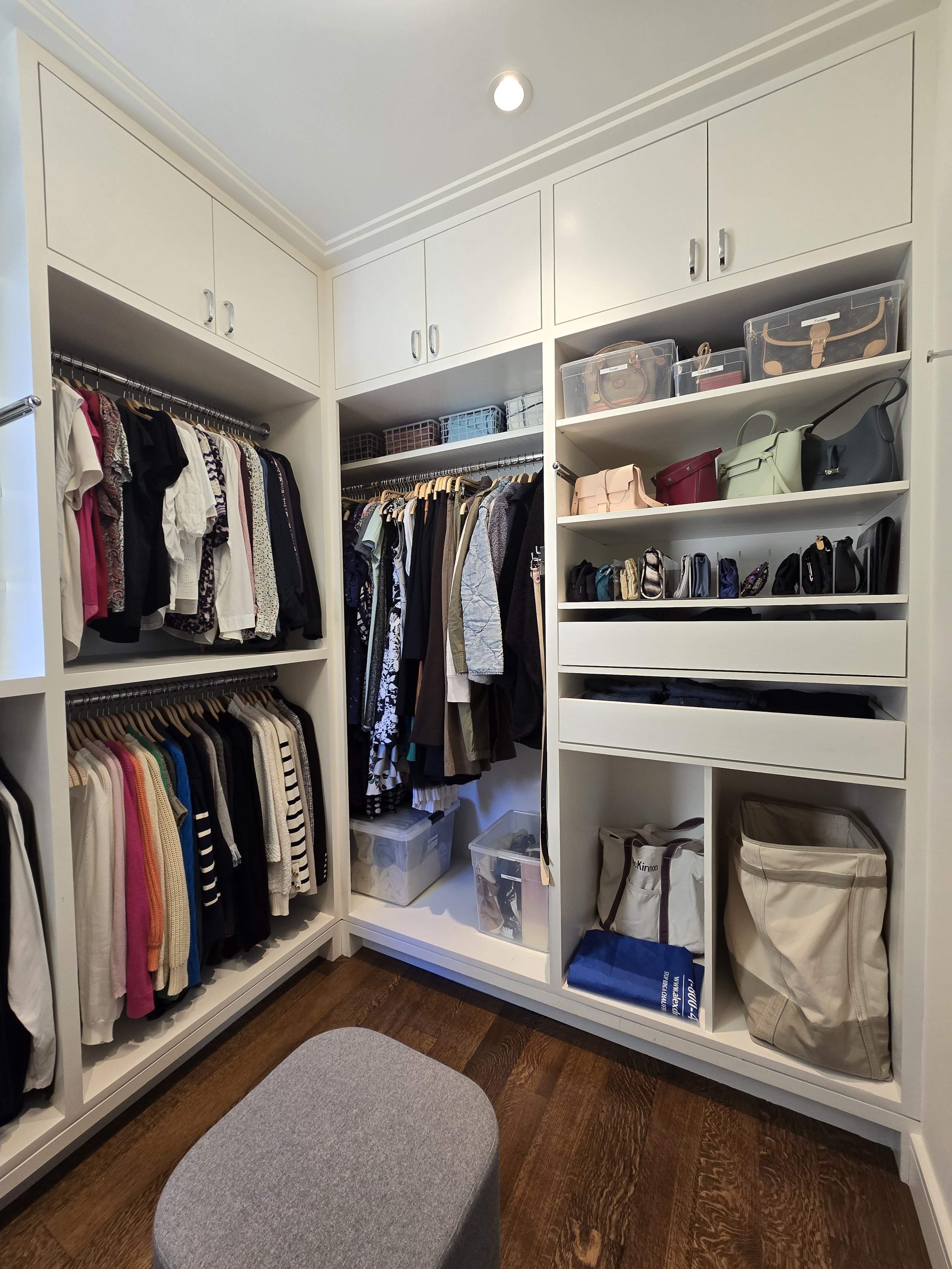 A walk-in closet with white built-in shelves and clothing hanging on racks, along with shelves holding handbags, boxes, and storage bins, and a gray ottoman on a dark wood floor.