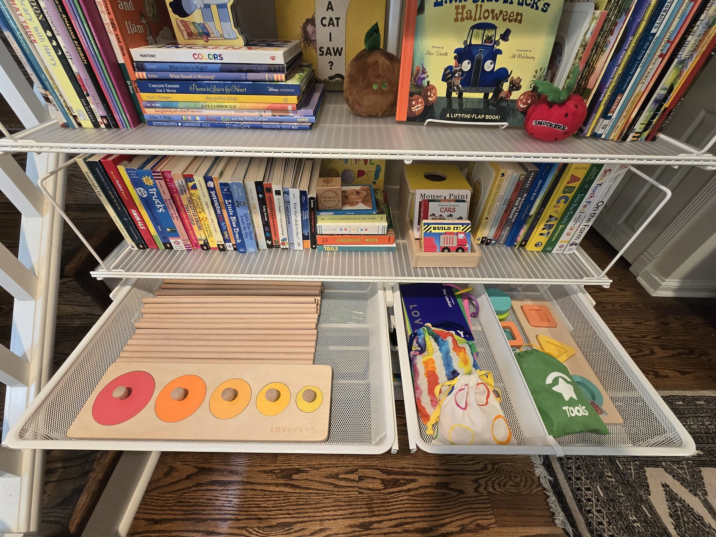 Bookshelves filled with children's books, toys, and educational tools. The top shelf has picture books, a small pumpkin decoration, and a plush apple. The middle shelves contain more children's books, including storybooks and activity books. The bott