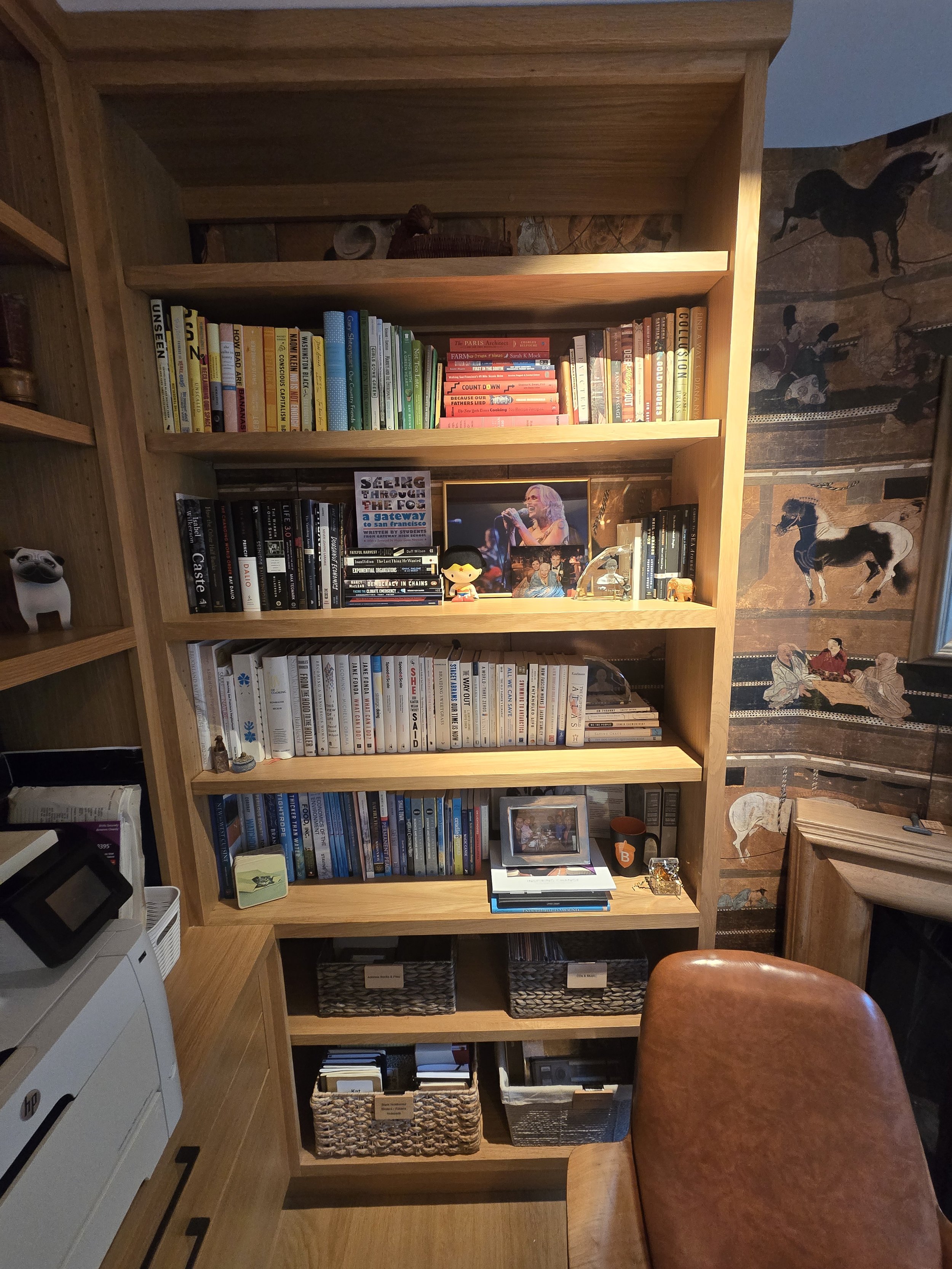 A wooden bookshelf filled with colorful books, framed photos, and decorative items, situated next to a wall with tribal art depicting animals and people, with a leather chair in front.