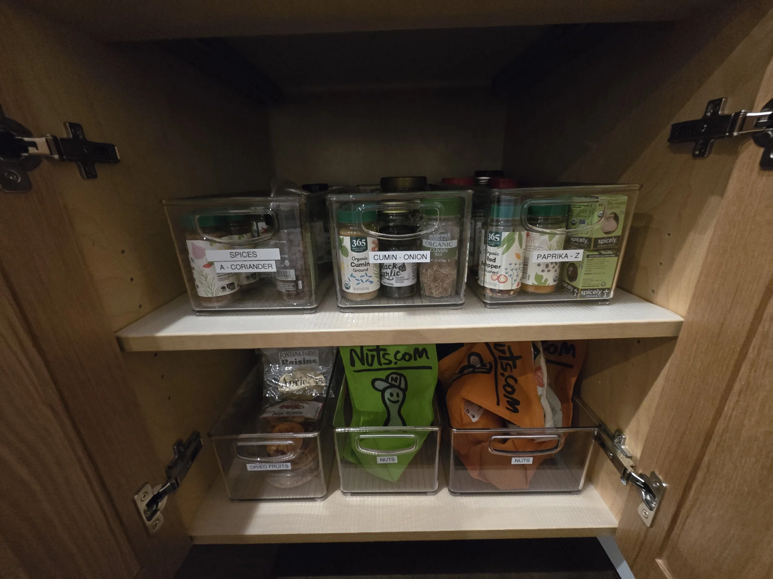 Shelf inside a wooden cabinet containing labeled spice jars on the top shelf and plastic containers with dried fruits and nuts on the bottom shelf.