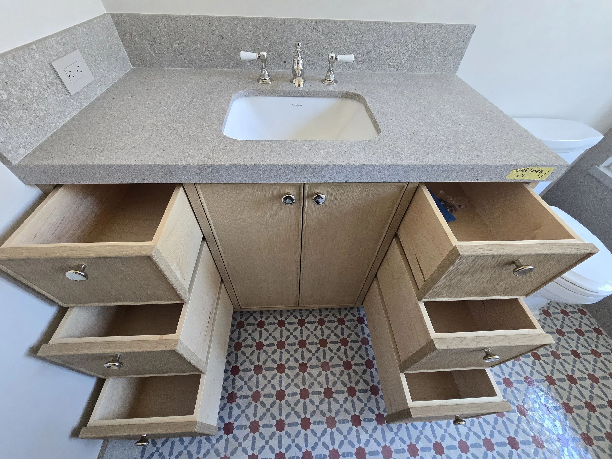 A bathroom vanity with an open drawer on the left, a closed drawer on the right, and a cabinet in the middle, topped with a gray speckled countertop and a built-in sink. There is a wall socket on the left side of the vanity and a toilet partially vis
