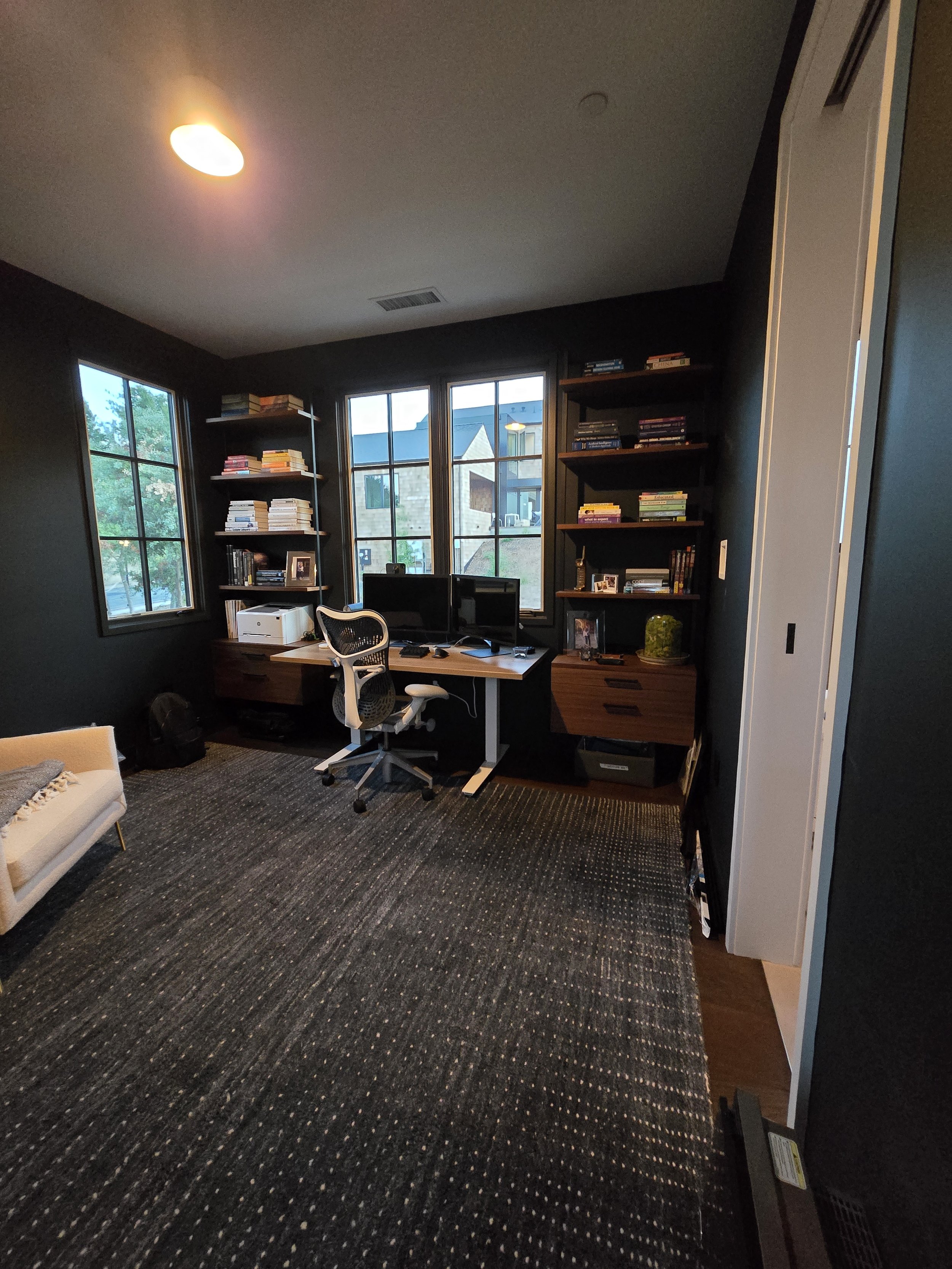 Home office with black walls, large windows, a desk with three monitors, a white ergonomic chair, bookshelves with books, and a beige sofa with a blanket.