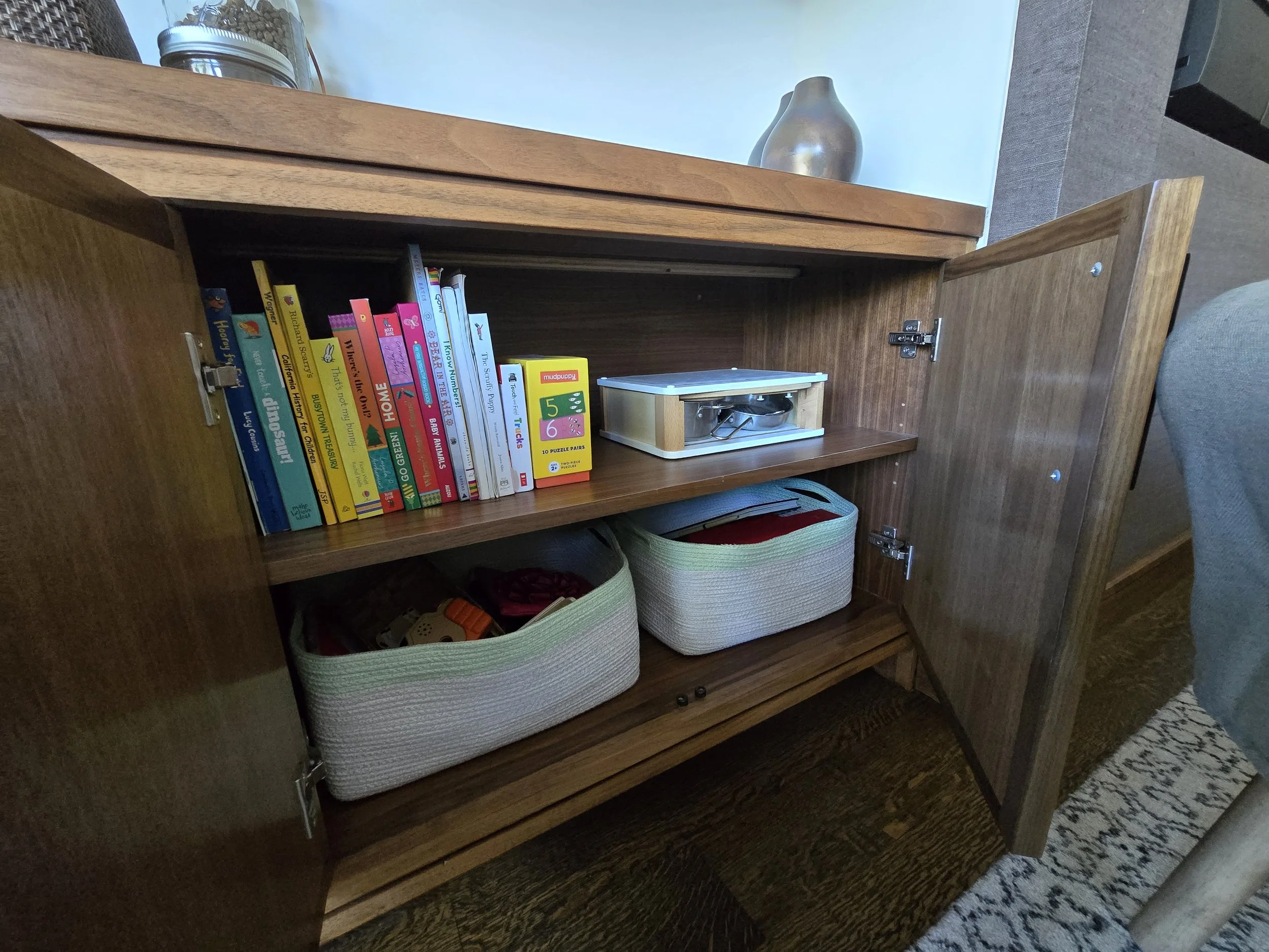 Open wooden cabinet with children’s books, a puzzle, a small metal tray, and fabric storage baskets inside.