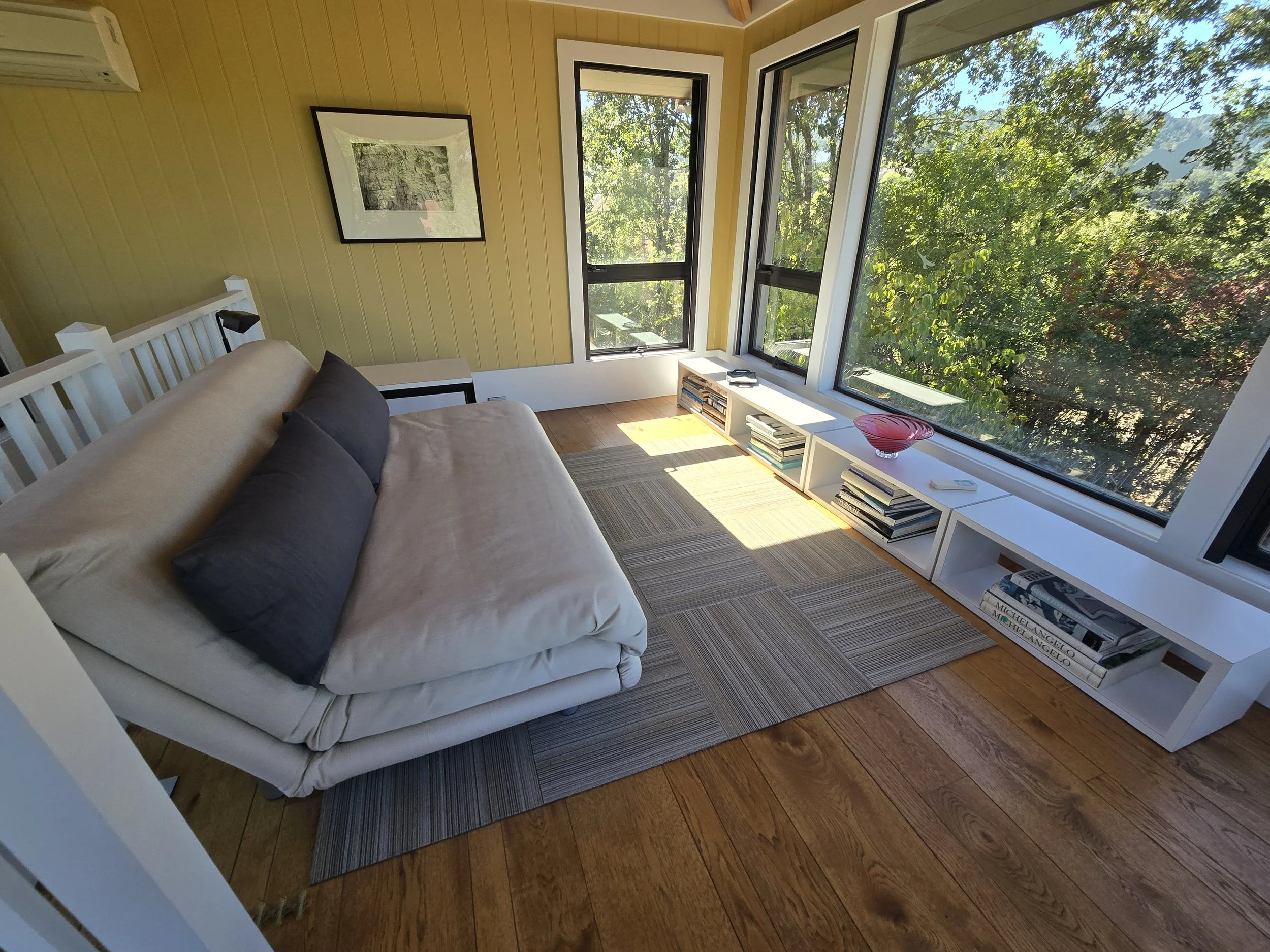 A living room with large windows showing trees outside, a white sofa with dark pillows, a striped rug, a white low bookshelf with books and a pink bowl, and a framed black-and-white artwork on a yellow wall.