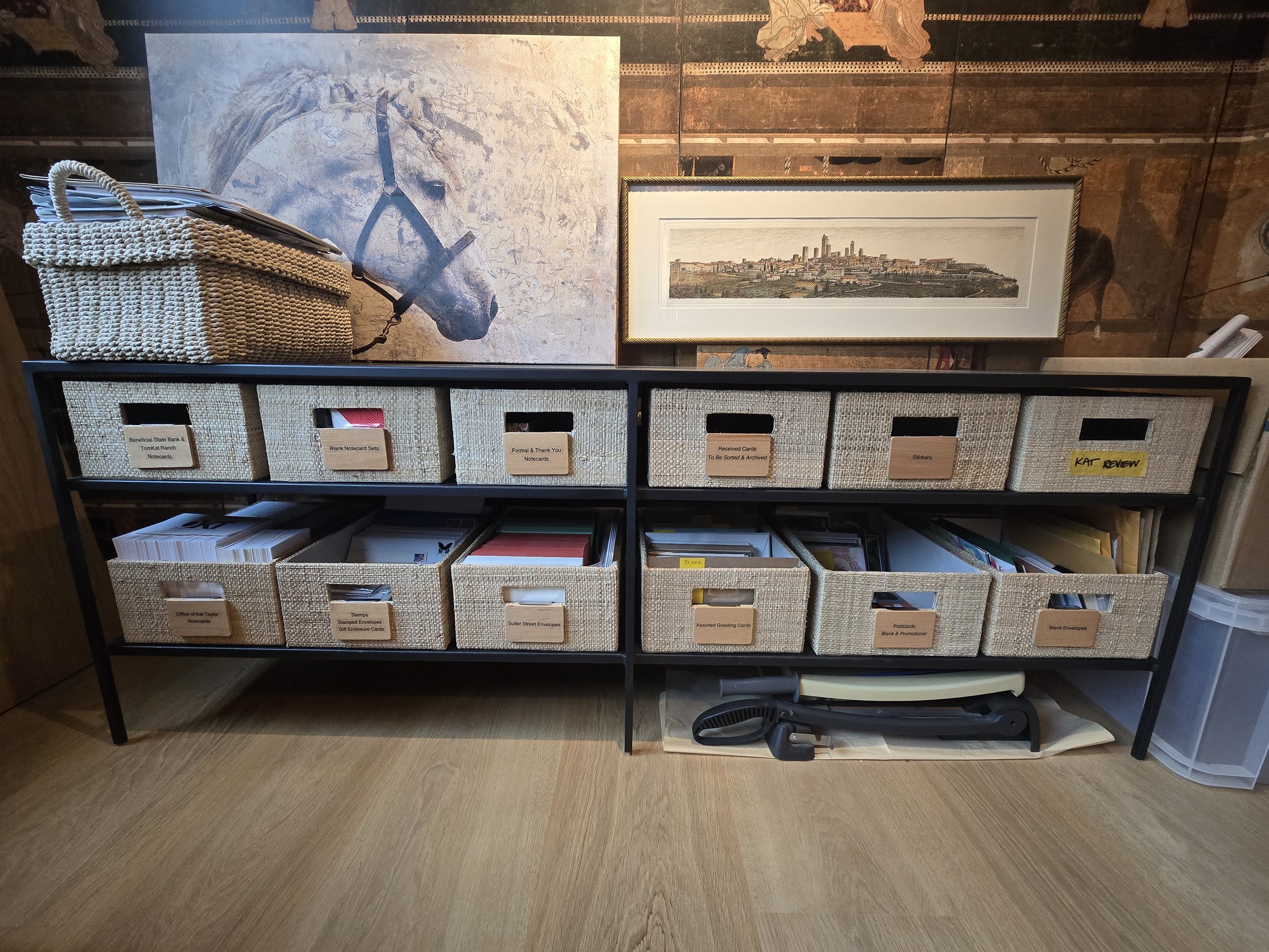 A black storage shelf with multiple woven baskets labeled for organizing letters, notecards, and documents, positioned in front of wall art and paintings.