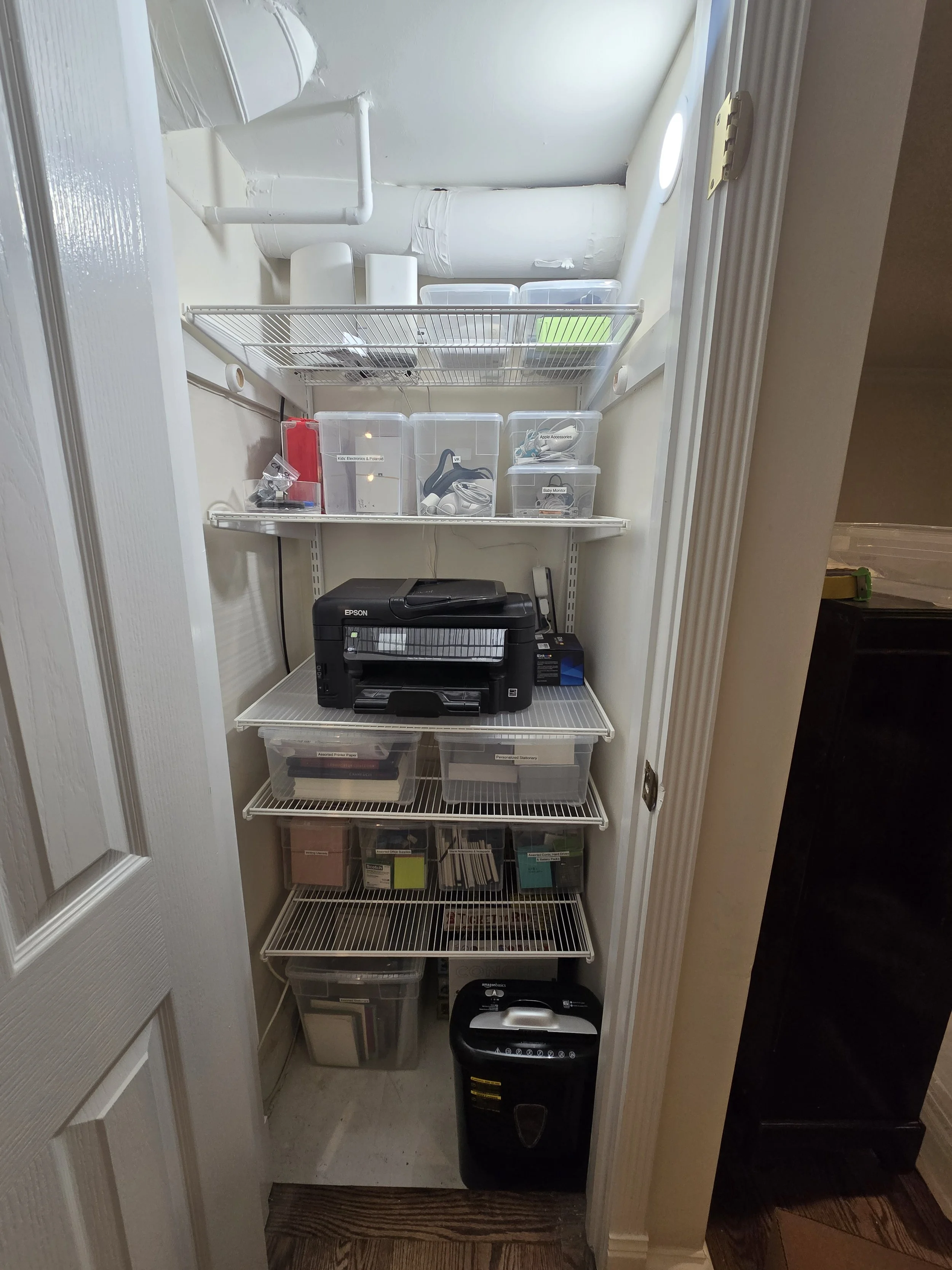A small walk-in storage closet with shelves holding various containers, a printer, and office supplies.