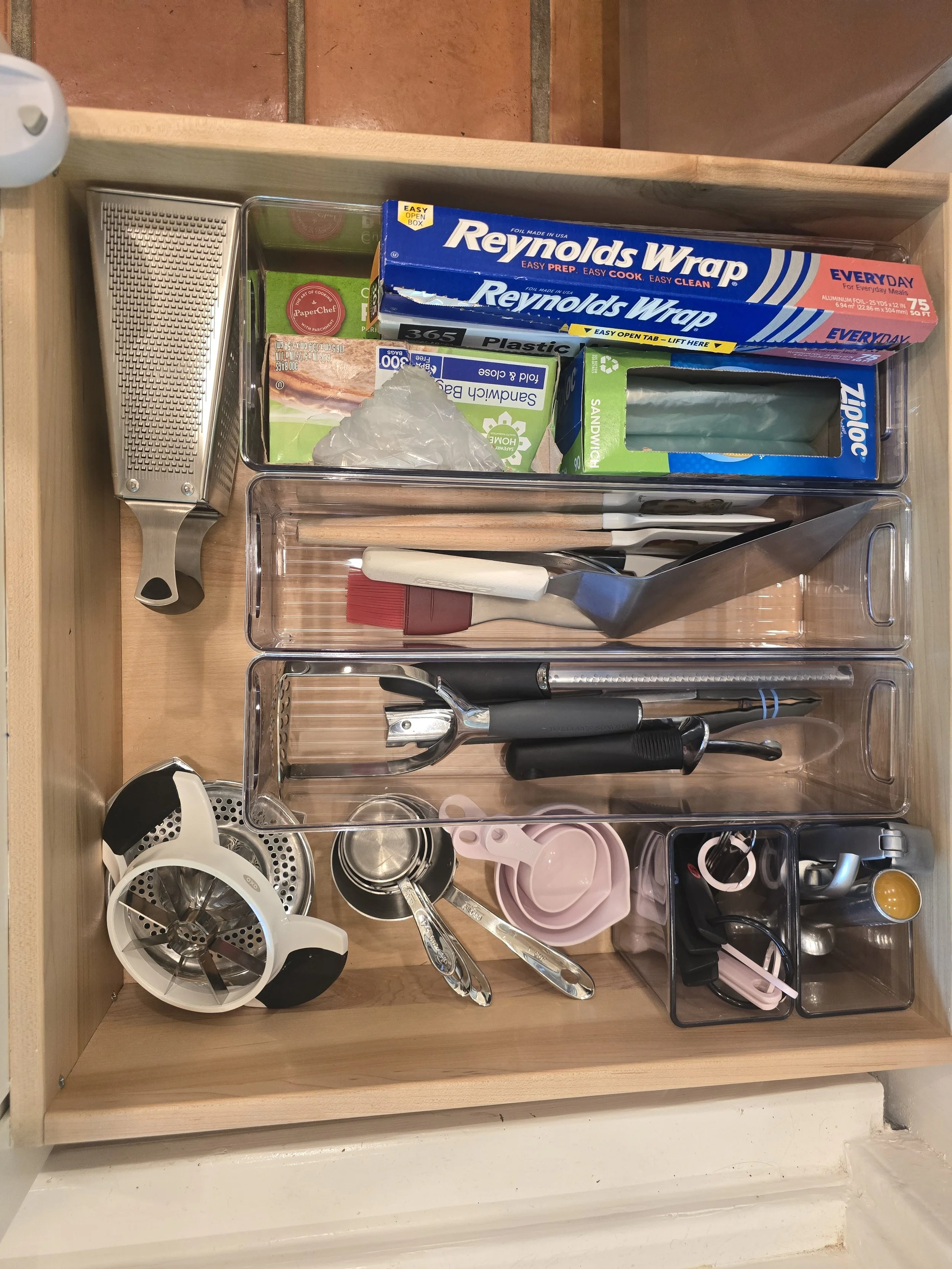 Drawers organized with kitchen utensils, plastic wrap, aluminum foil, sandwich bags, measuring cups, milk frother, small bowls, measuring spoons, and a knife.
