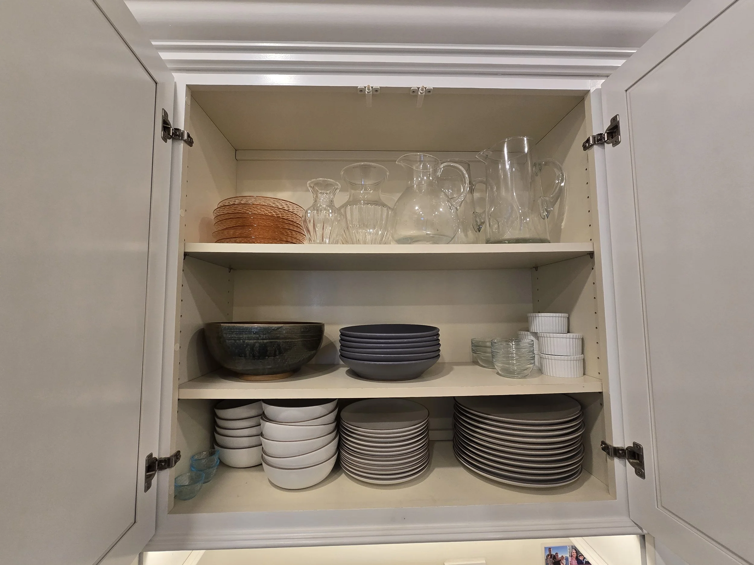Open kitchen cabinet with glass pitchers, bowls, plates, and small glass cups on three shelves.