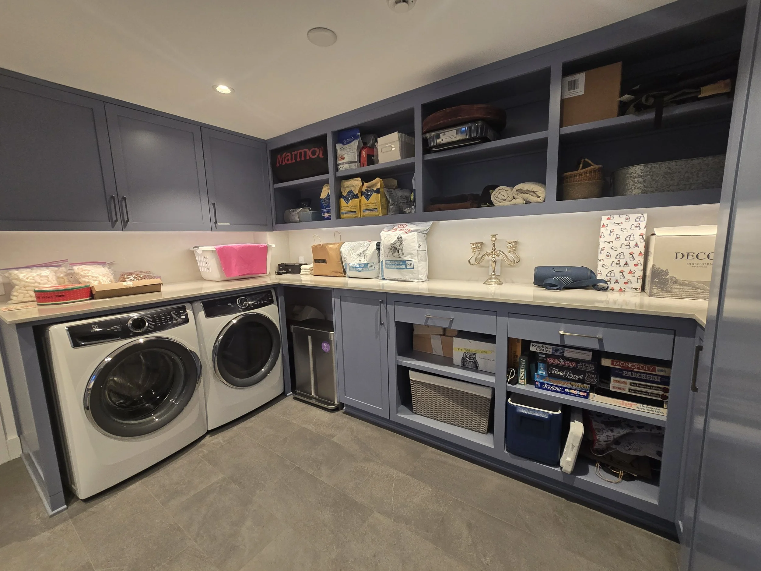 A laundry room with a front-loading washing machine and dryer, dark blue cabinetry, open shelves with various items, a countertop with laundry supplies, and a tiled floor.