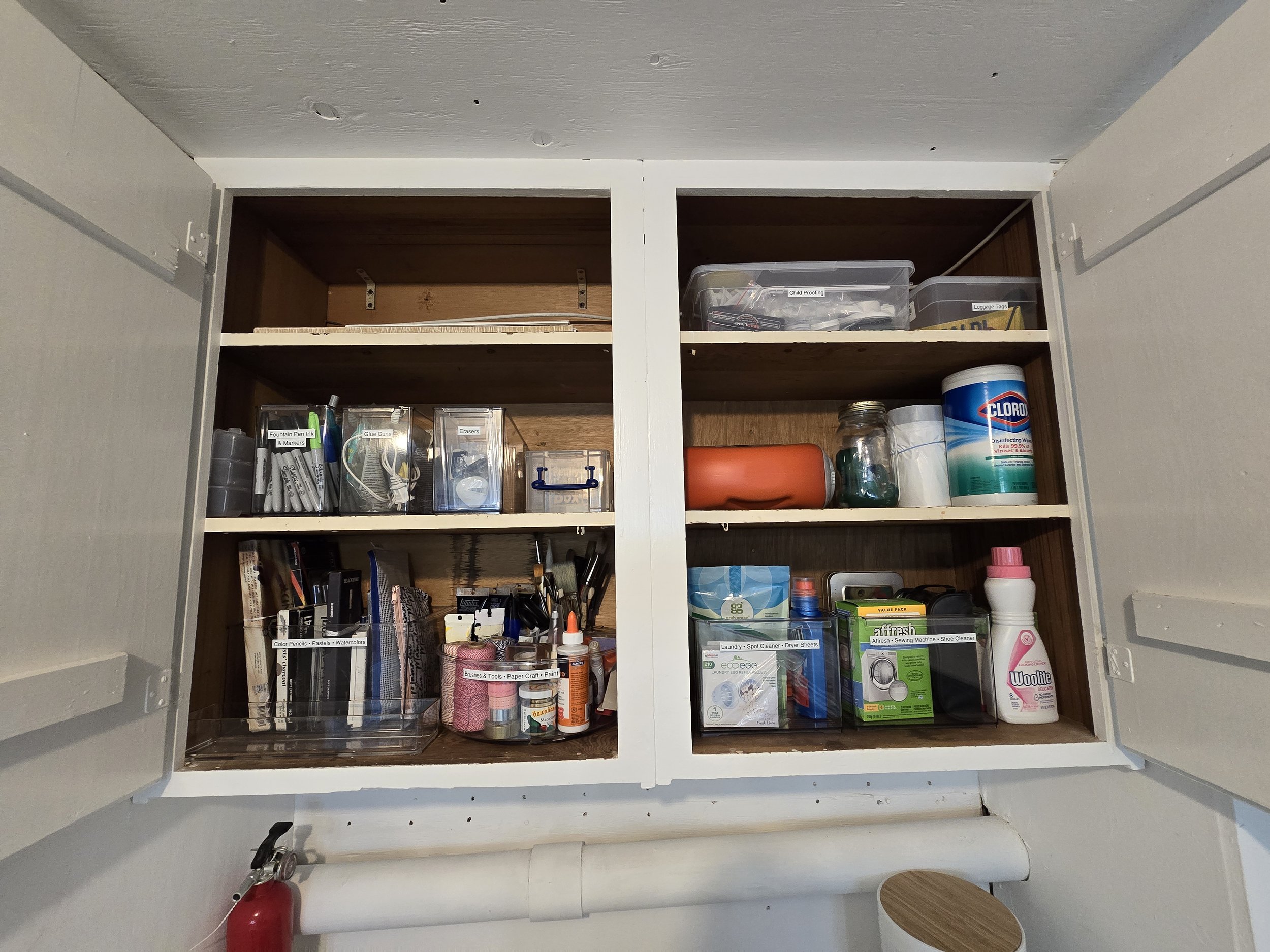 Open kitchen cabinet with two shelves filled with office supplies, cleaning products, and miscellaneous items.